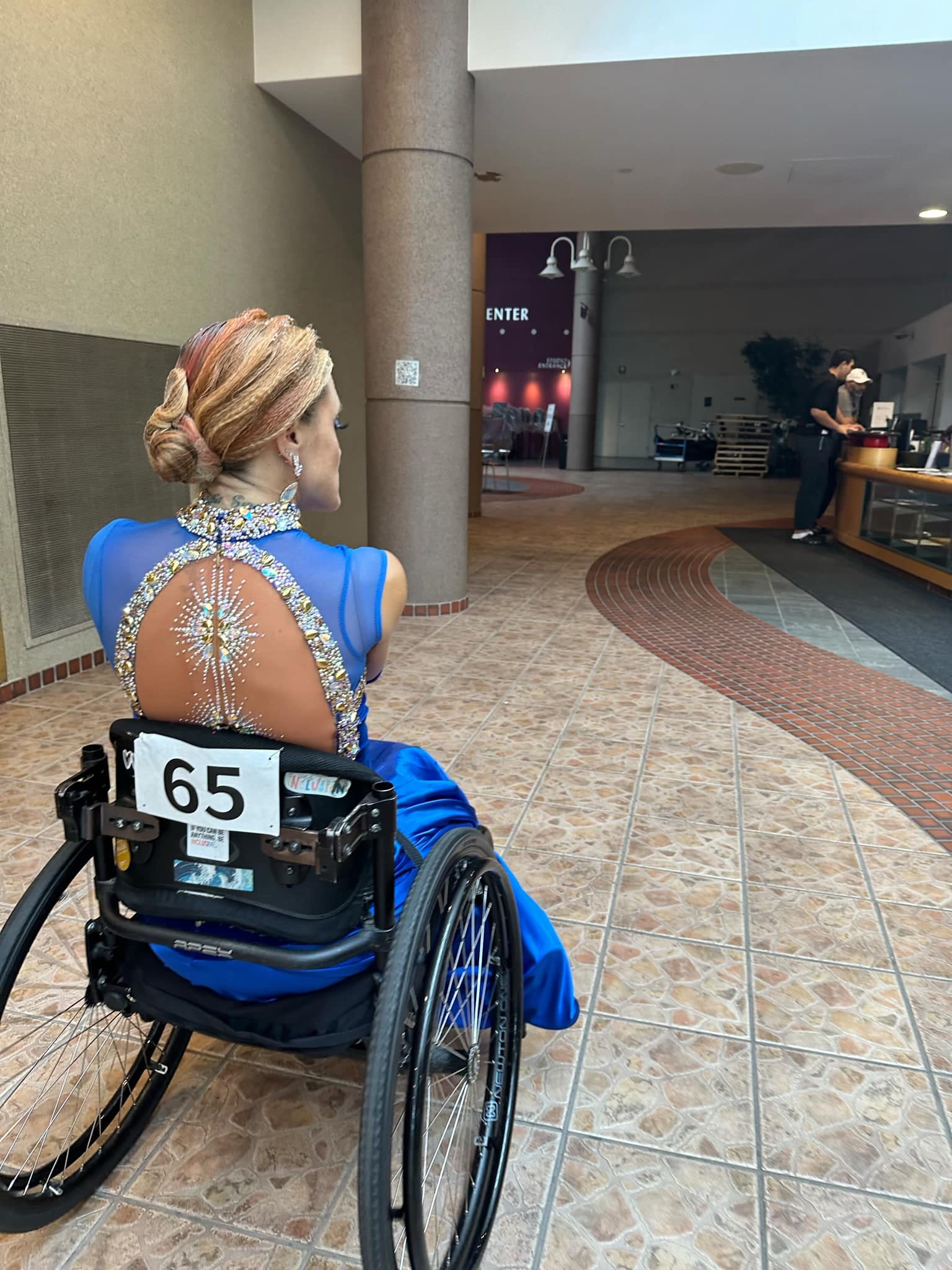 A woman with pink and orange streaks in her hair and a blue sequin dress sits in a wheelchair with her back to the camera