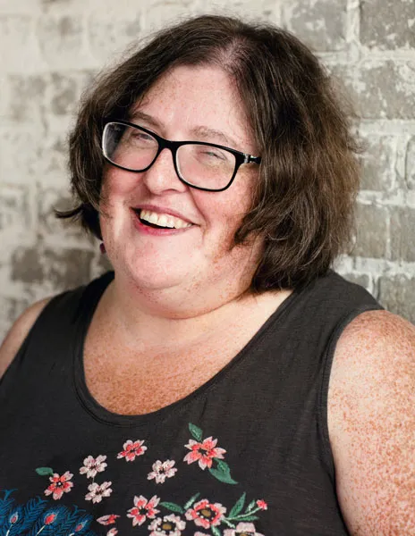 A woman with short brown hair and glasses is smiling while wearing a black tank top with embroidered flowers