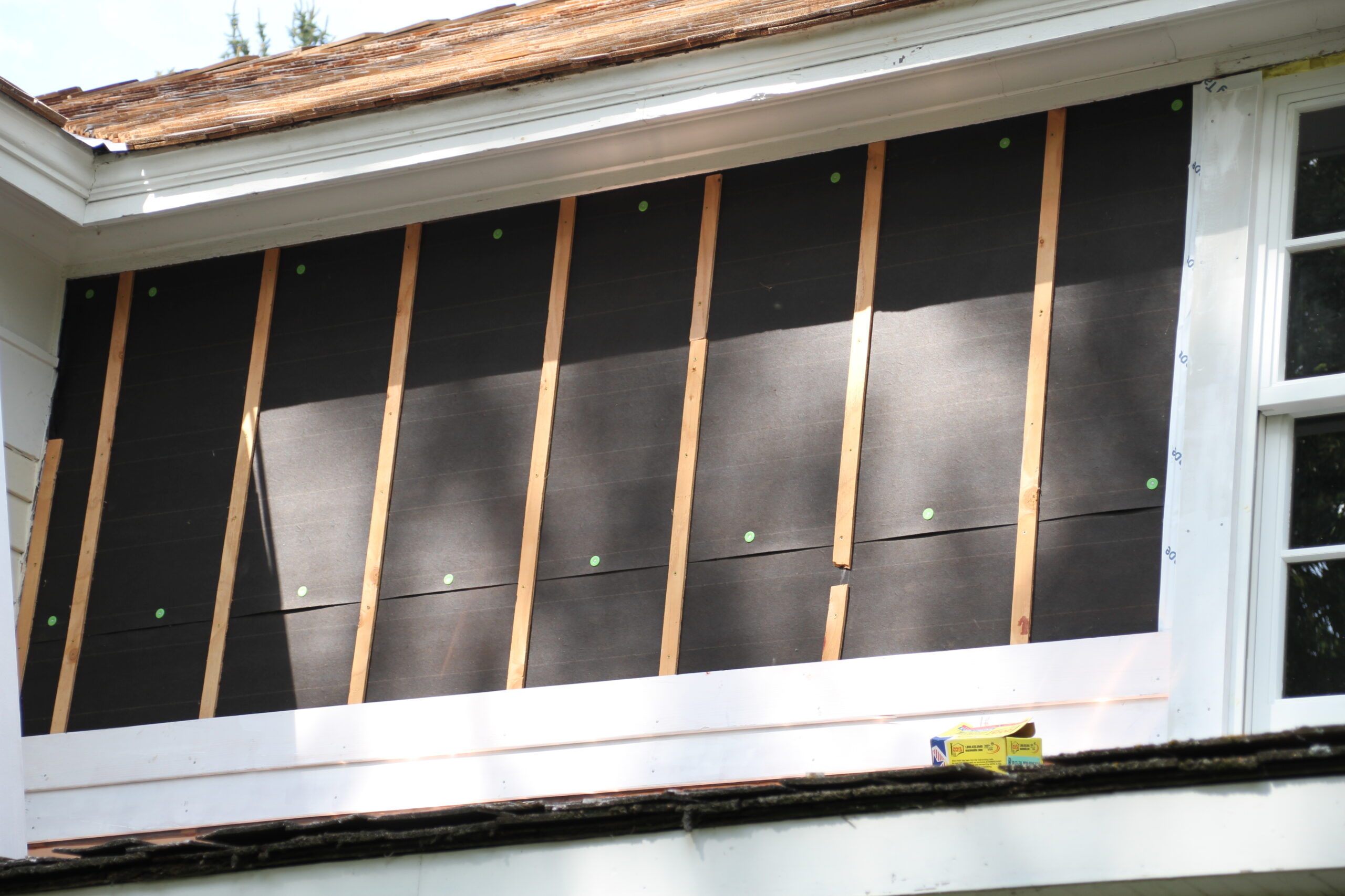 An exterior wall of a home is covered with black tar paper. Over the tar paper, there are plywood strips. The home is an older one --- it was built in 1861.