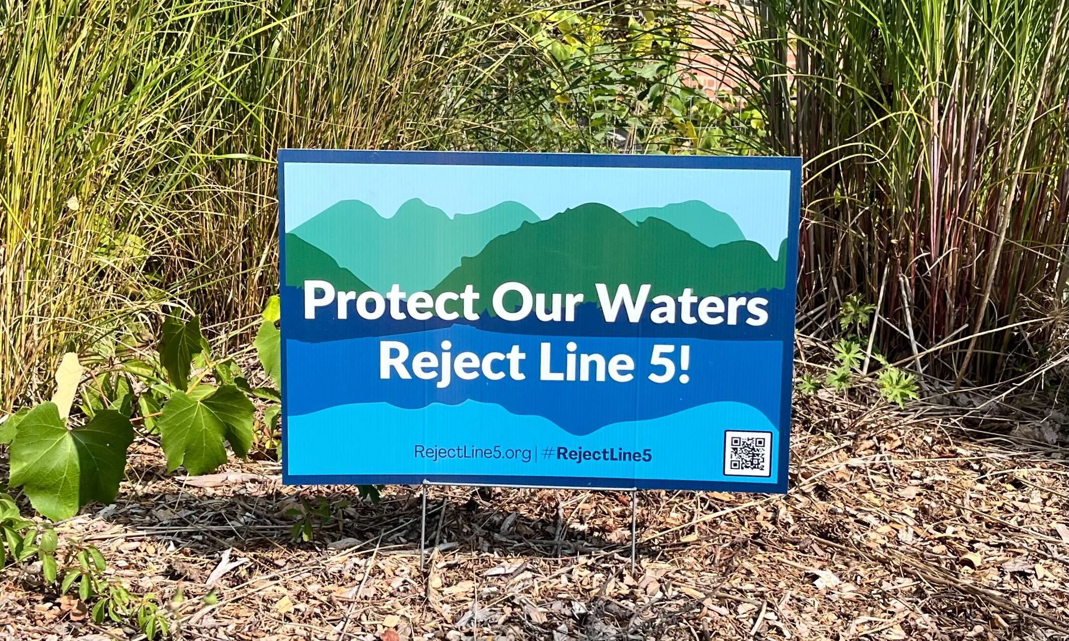 A sign with green and blue natural artwork says "Protect Our Waters Reject Line 5!"