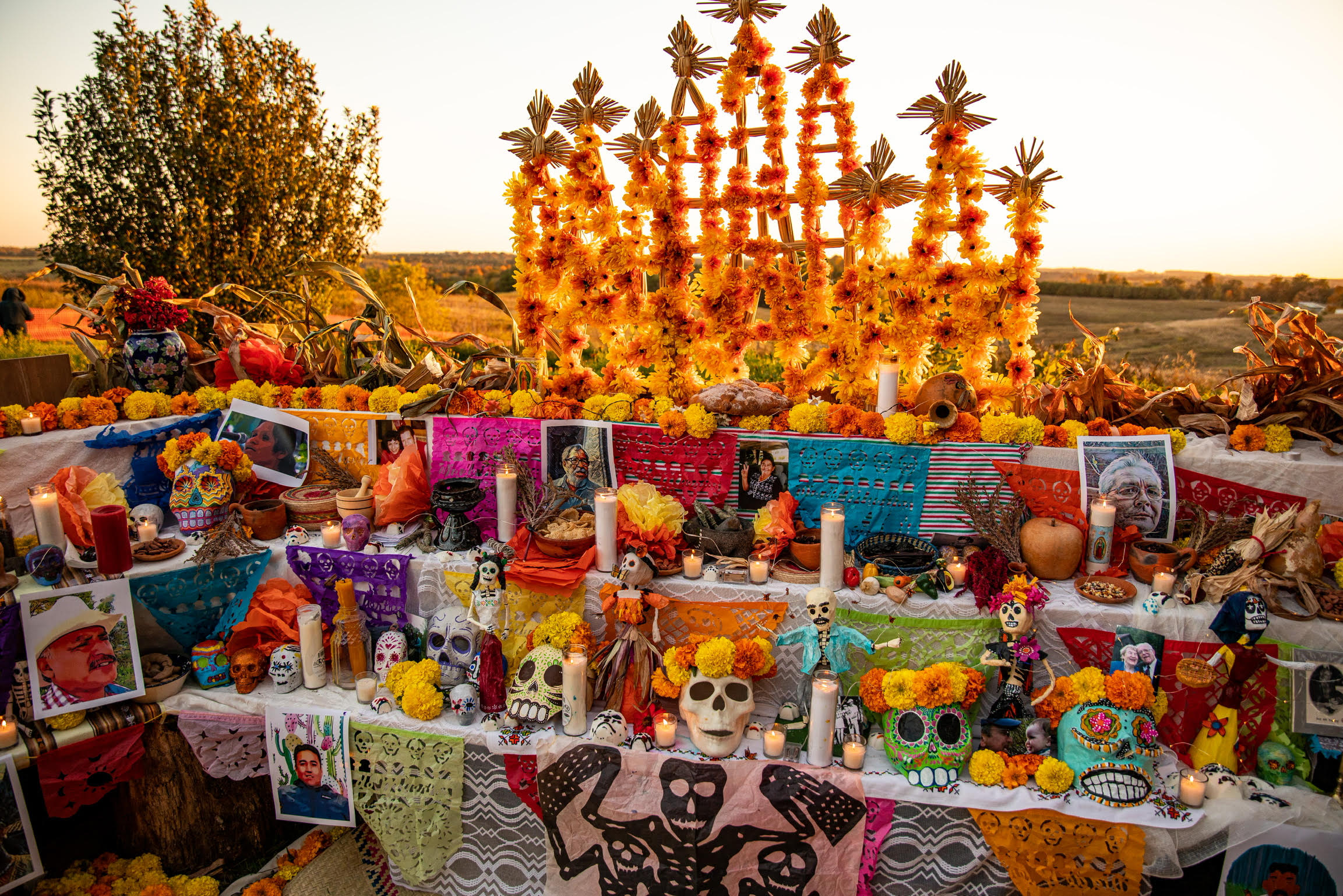 A colorful array of banners, skulls, flowers, and photos set against a rolling farmland backdrop on a sunny day