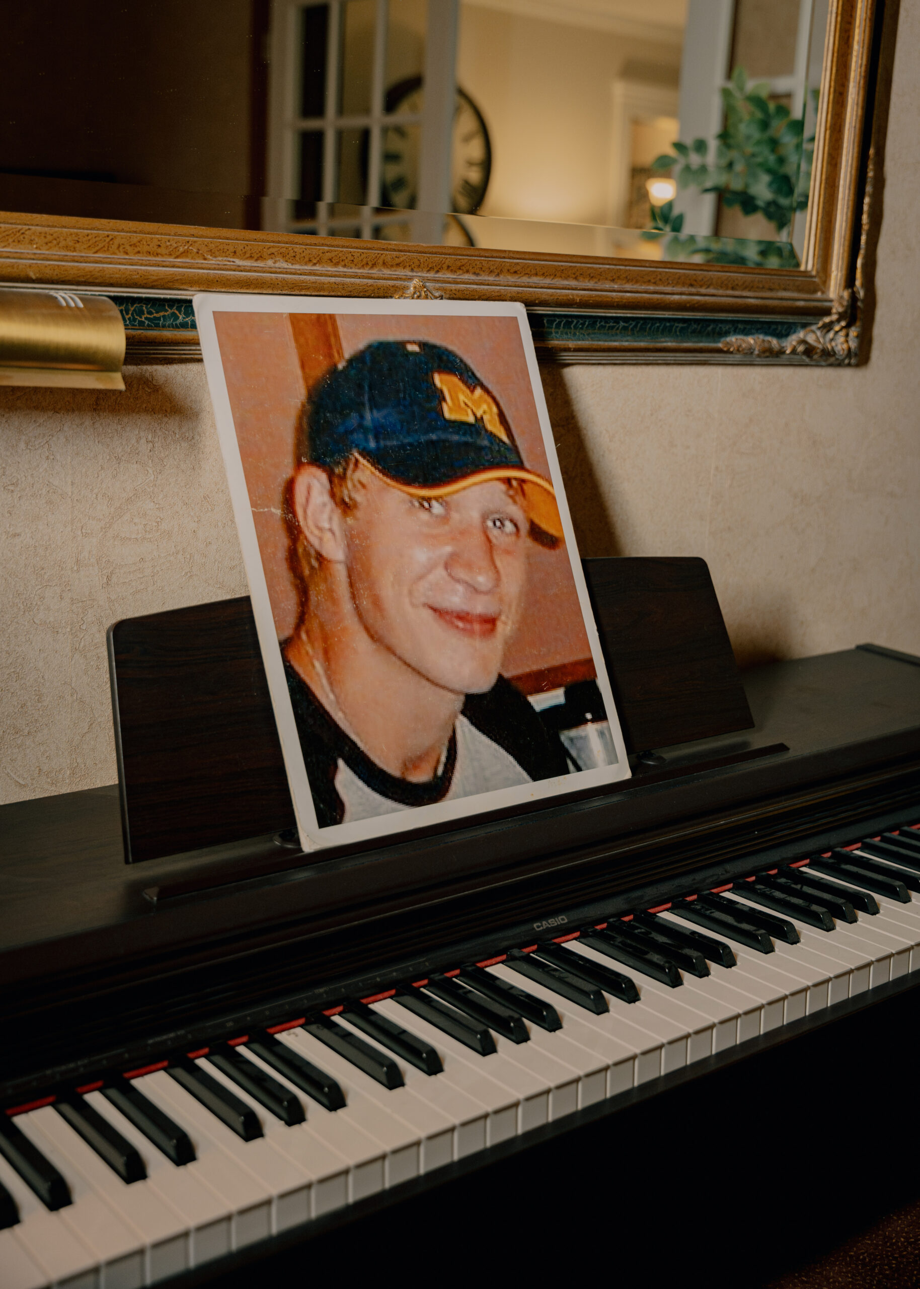 A photo of Michael Bell Jr., a White man, wearing a blue hat with a yellow "M" on it, sits on a piano.