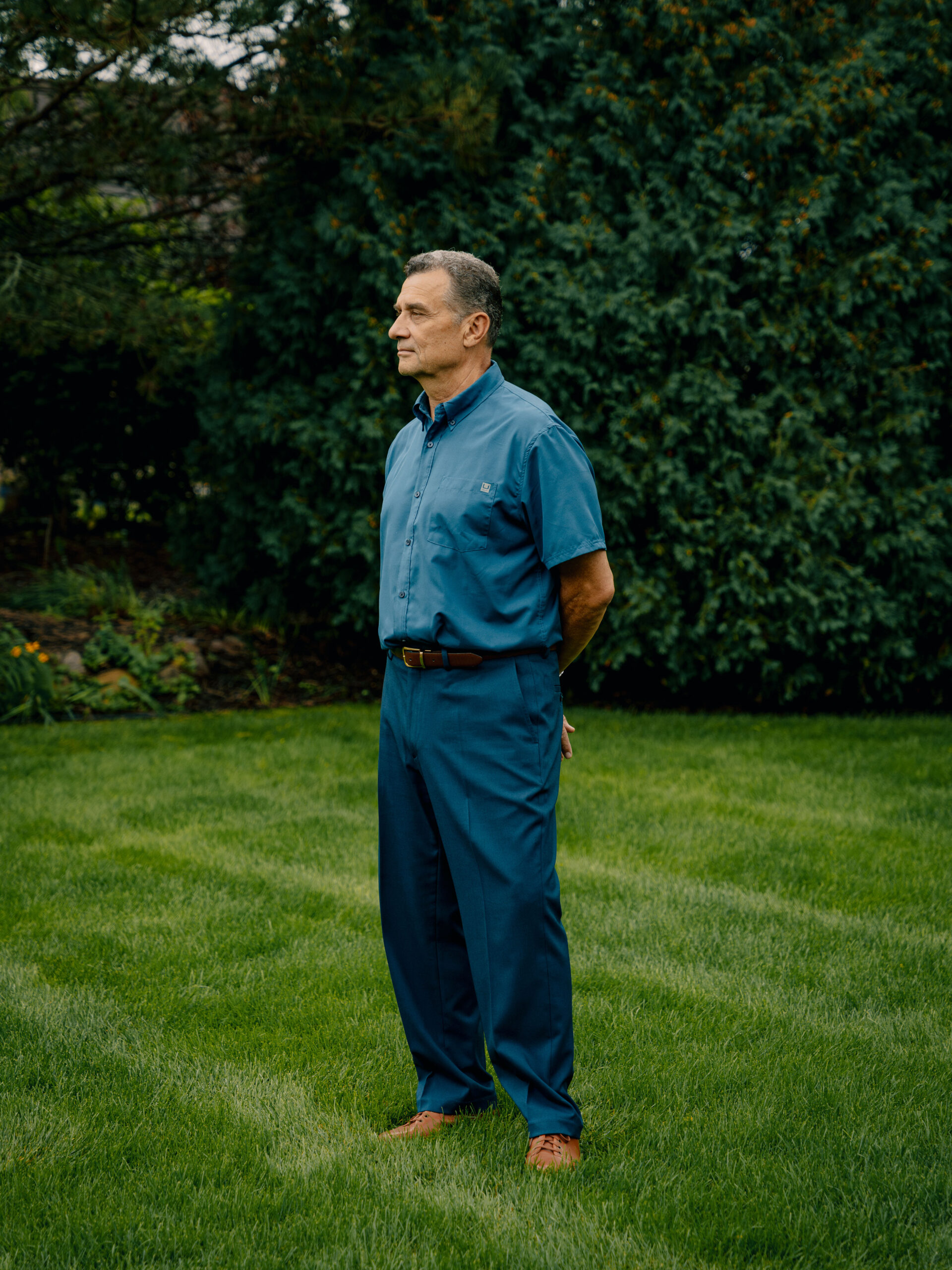 Michael Bell Sr., a White man with gray hair, wears a blue short-sleeved shirt and blue pants. He stands in a yard with green grass and trees.