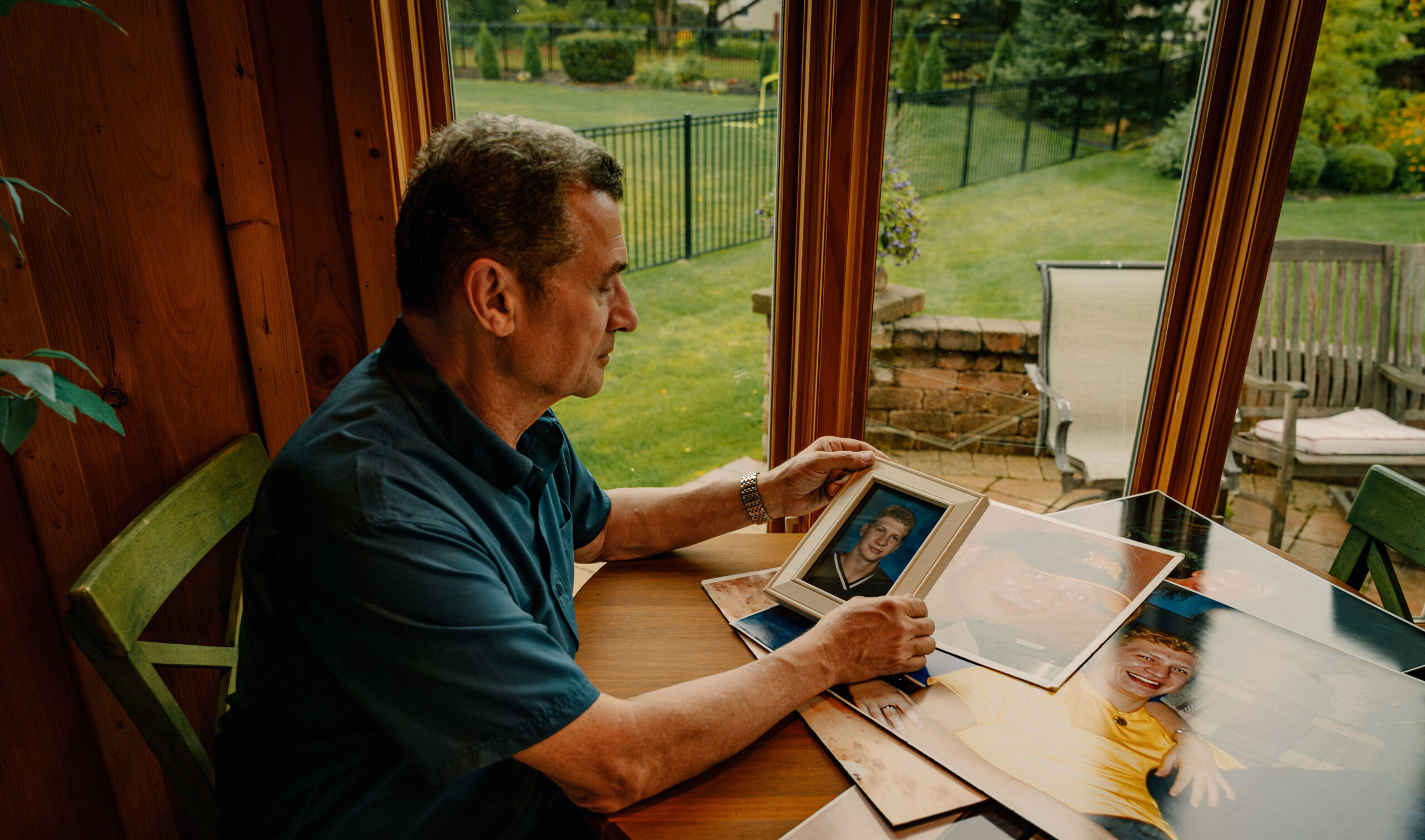 Michael Bell Sr., a White man, looks at a high school photo of his son, Michael Bell Jr., a White man, while sitting at a table. Four other photos of Michael are visible beneath the high school photo of Michael Bell Jr.