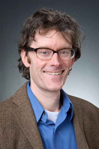 A middle-aged white man with glasses and a brown suit jacked smiles for a professor photo.