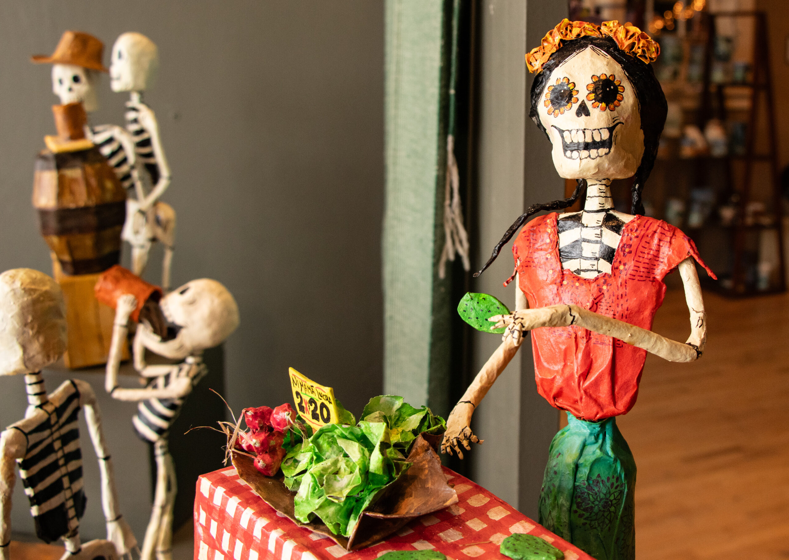 A feminine skeleton wearing a flower crown stands behind a checkered tablecloth selling cardboard lettuce and radishes