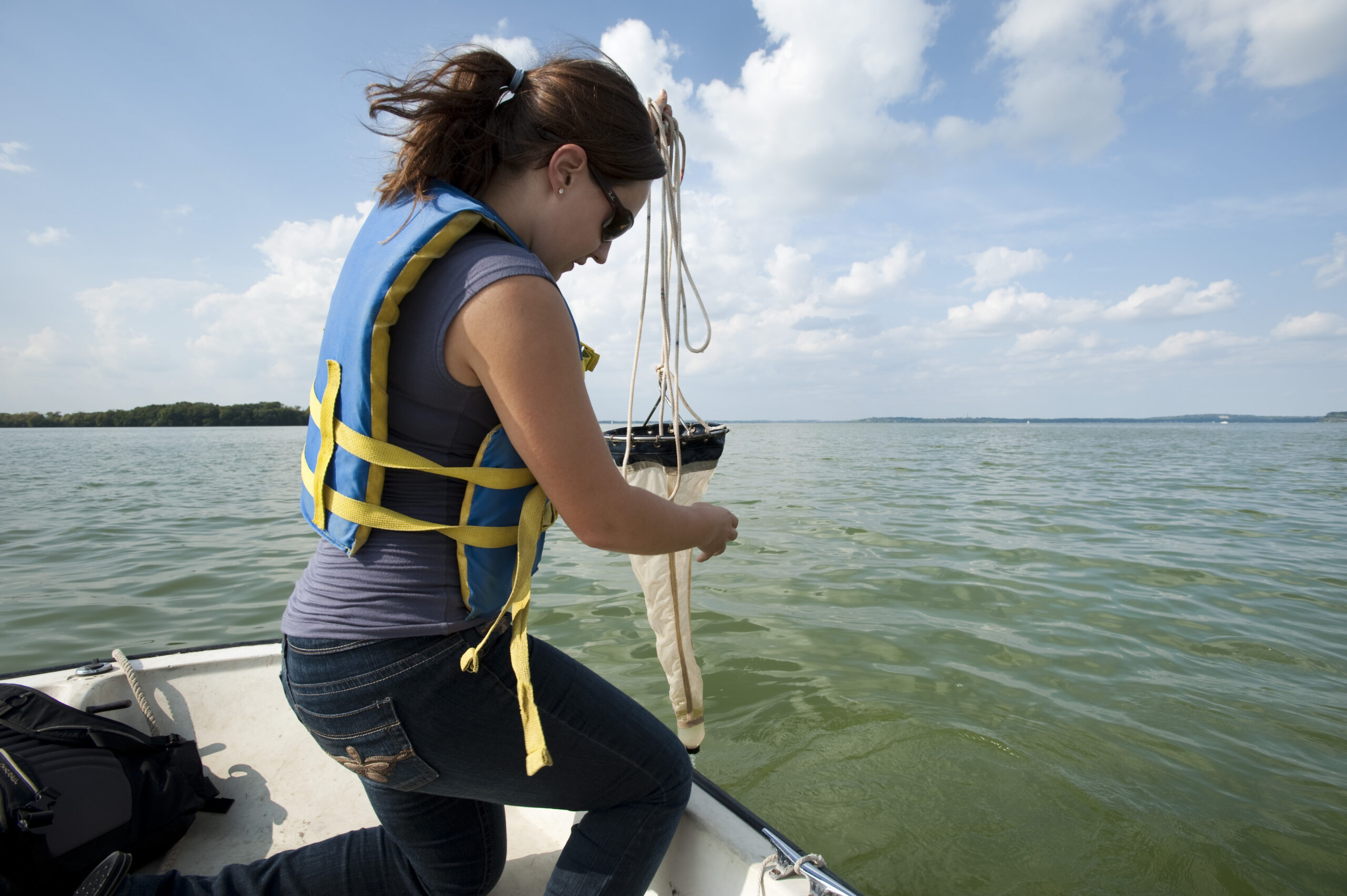 A young woman wearing a life jacket kneels on a boat and pulls a net out of a lake.