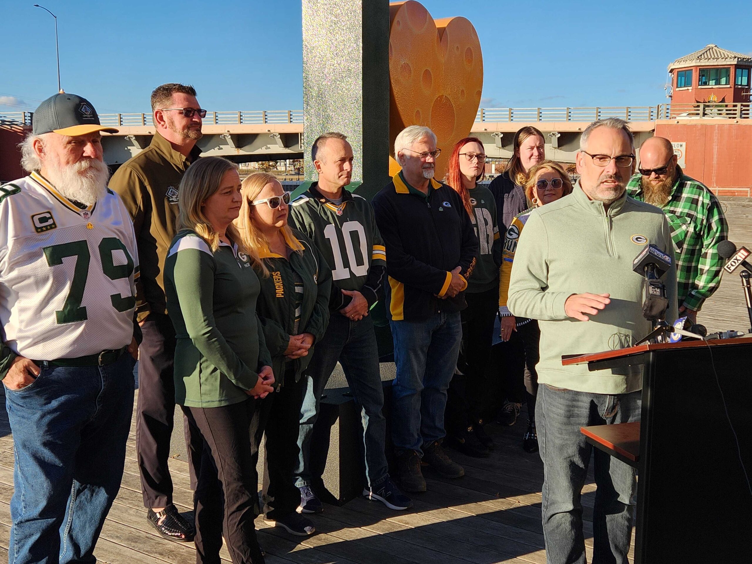 Green Bay City Council President Brian Johnson speaks at a podium as the other 11 city council members stand behind him on Wednesday, Oct. 23, 2024. The council held a press conference to stand behind the mayor in Lambeau Field lease negotiations between the Green Bay Packers and the city.