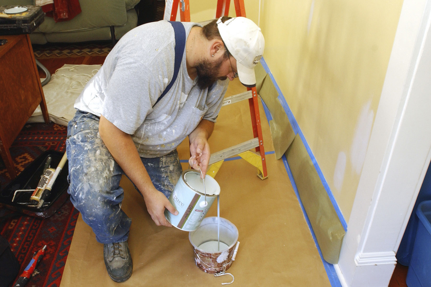 A painter poors white paint into another bucket. He's taped the wall in front of him to ready it for painting.