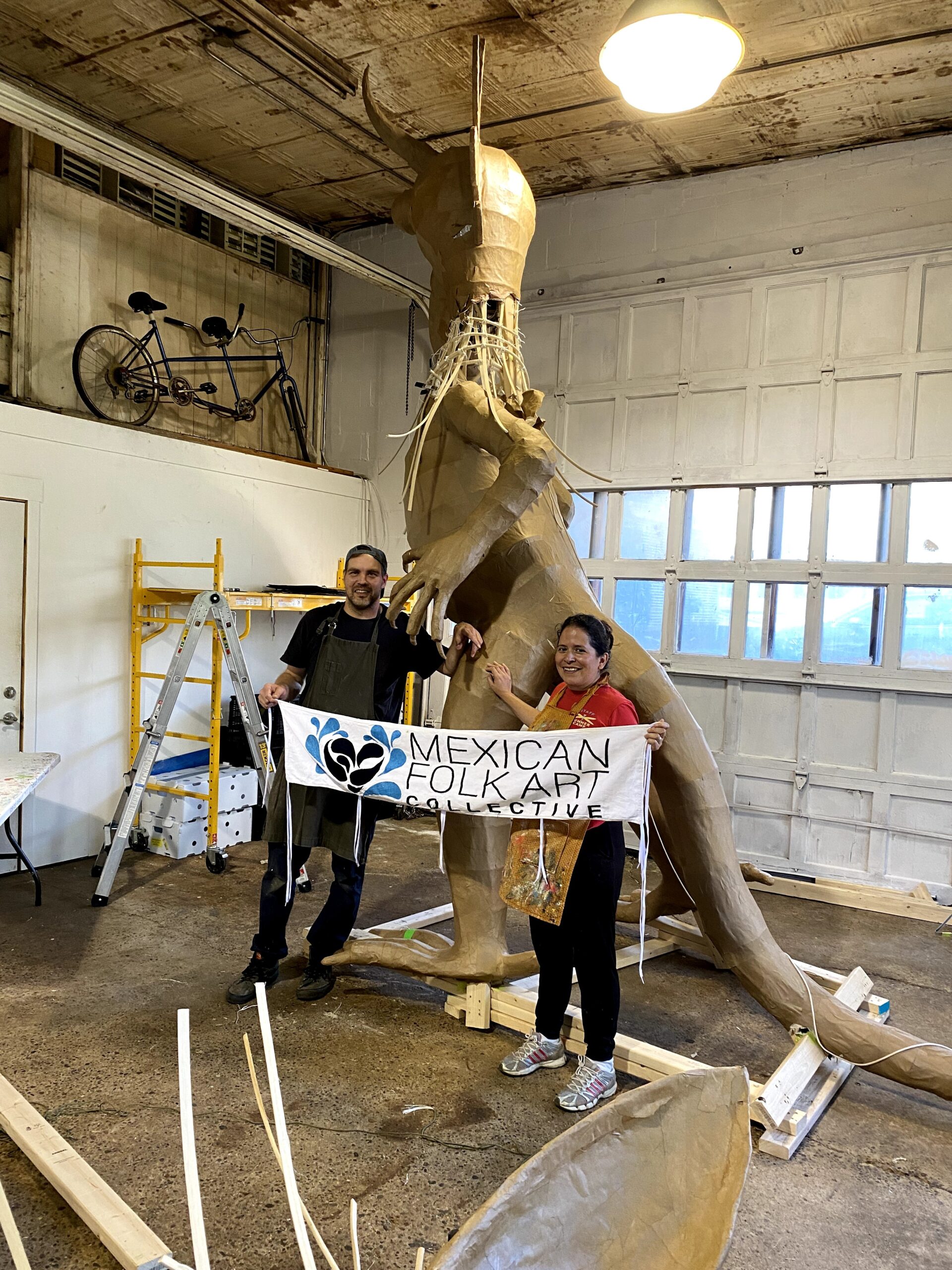 A man and a woman in aprons stand next to a tall statue of a cardboard creature holding a sign reading "Meixcan Folk Art Collective"