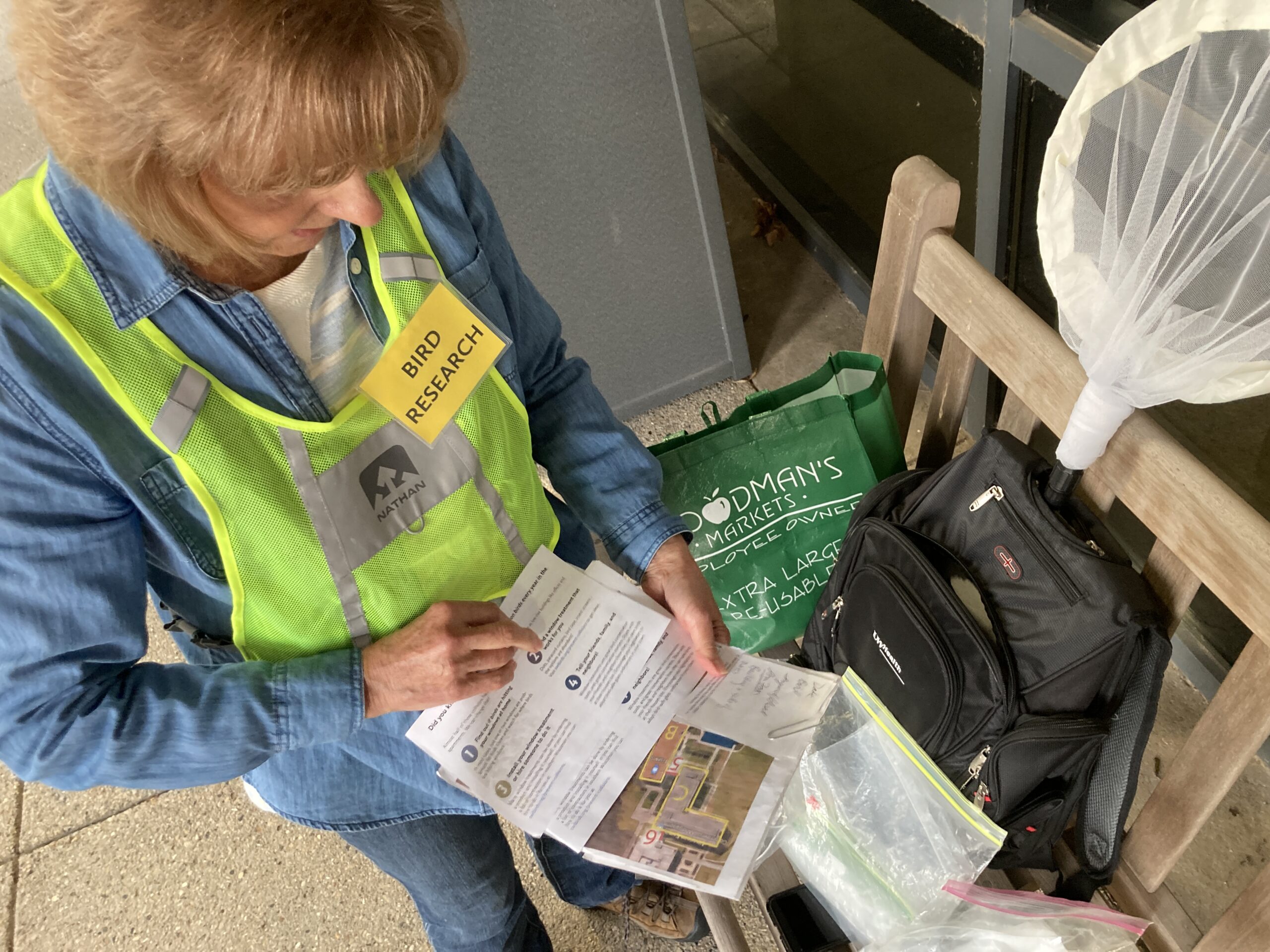 Volunteer Ann Mader wears a bright vest and gathers materials to survey birds. She holds a map of the site, and has a backpack, canvas bag and net beside her.