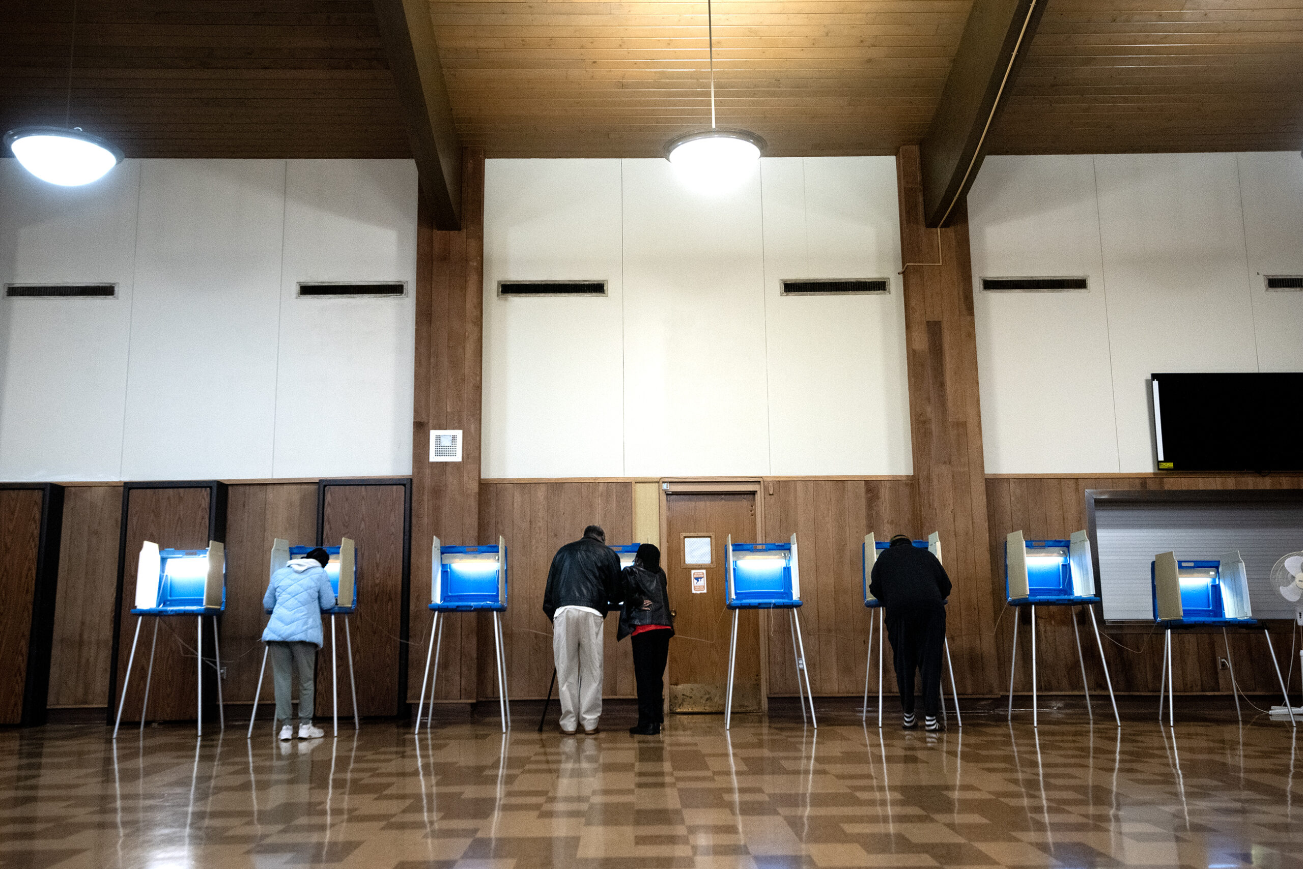 People standing at voting booths in a large room with wooden walls and a tiled floor.