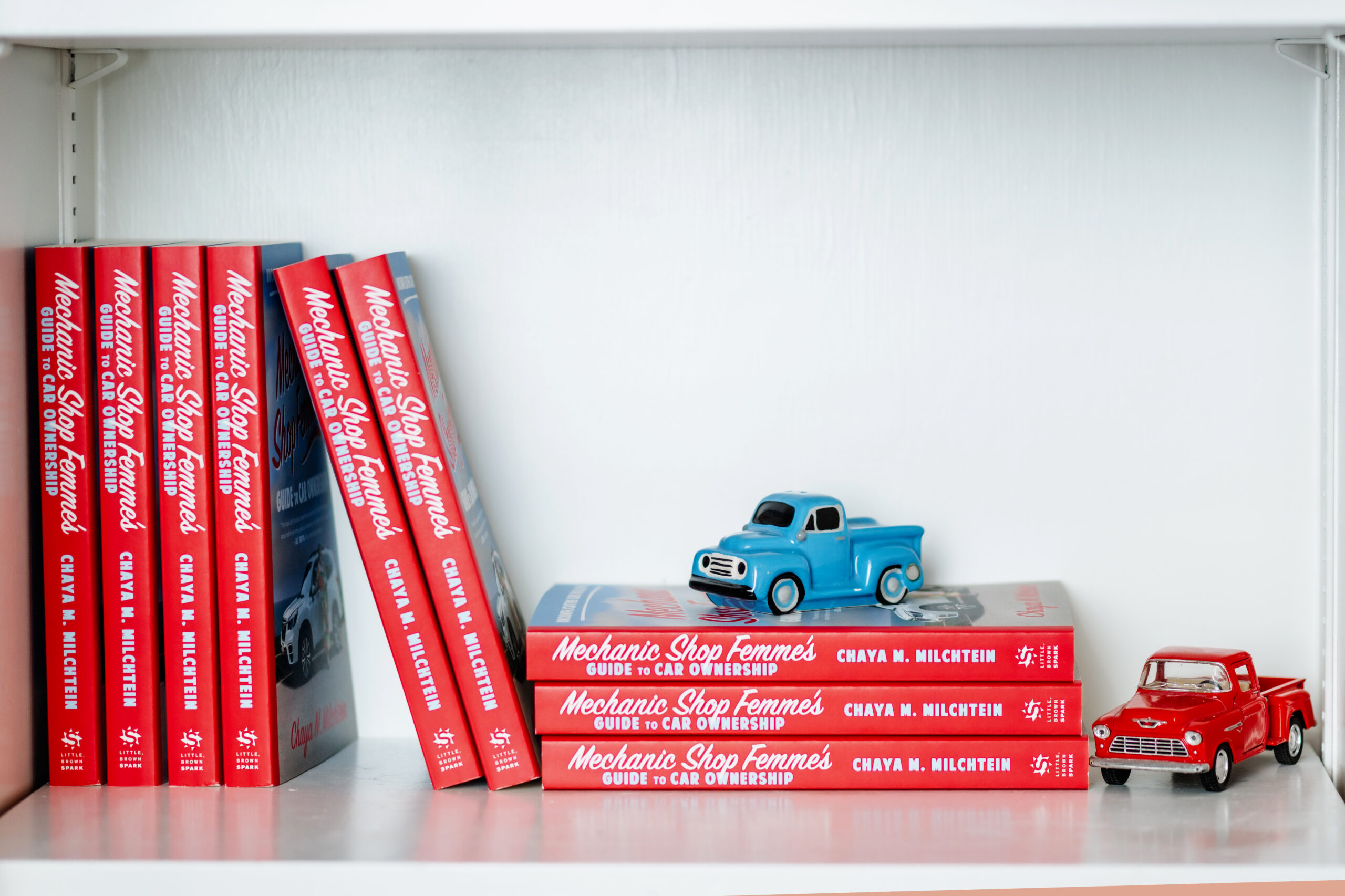 Books with a bright red spine are displayed on a metal bookshelf, with vintage car figurines that are blue and red