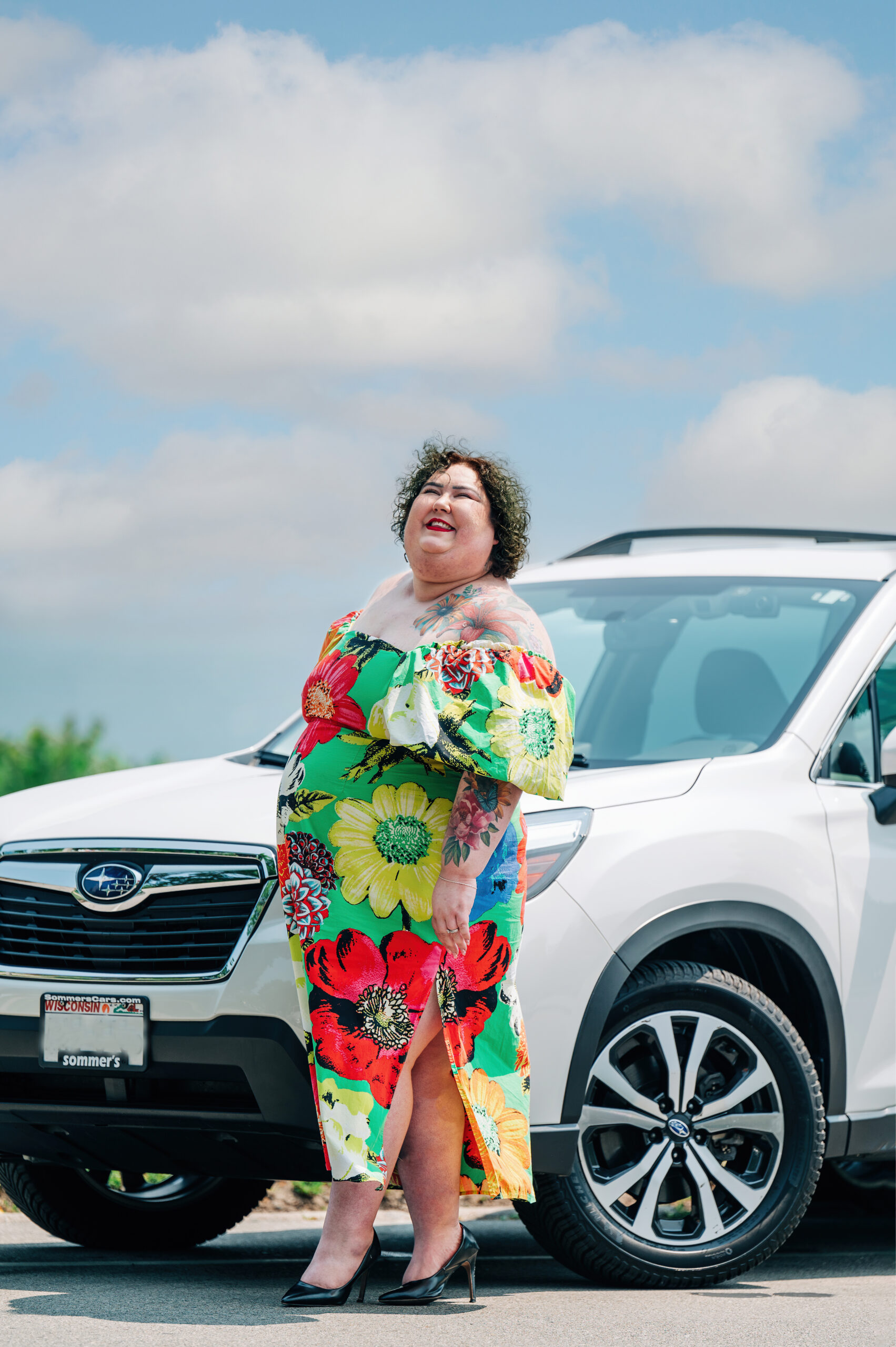 A woman in a bright floral dress and heels stands in front of a white Subaru SUV