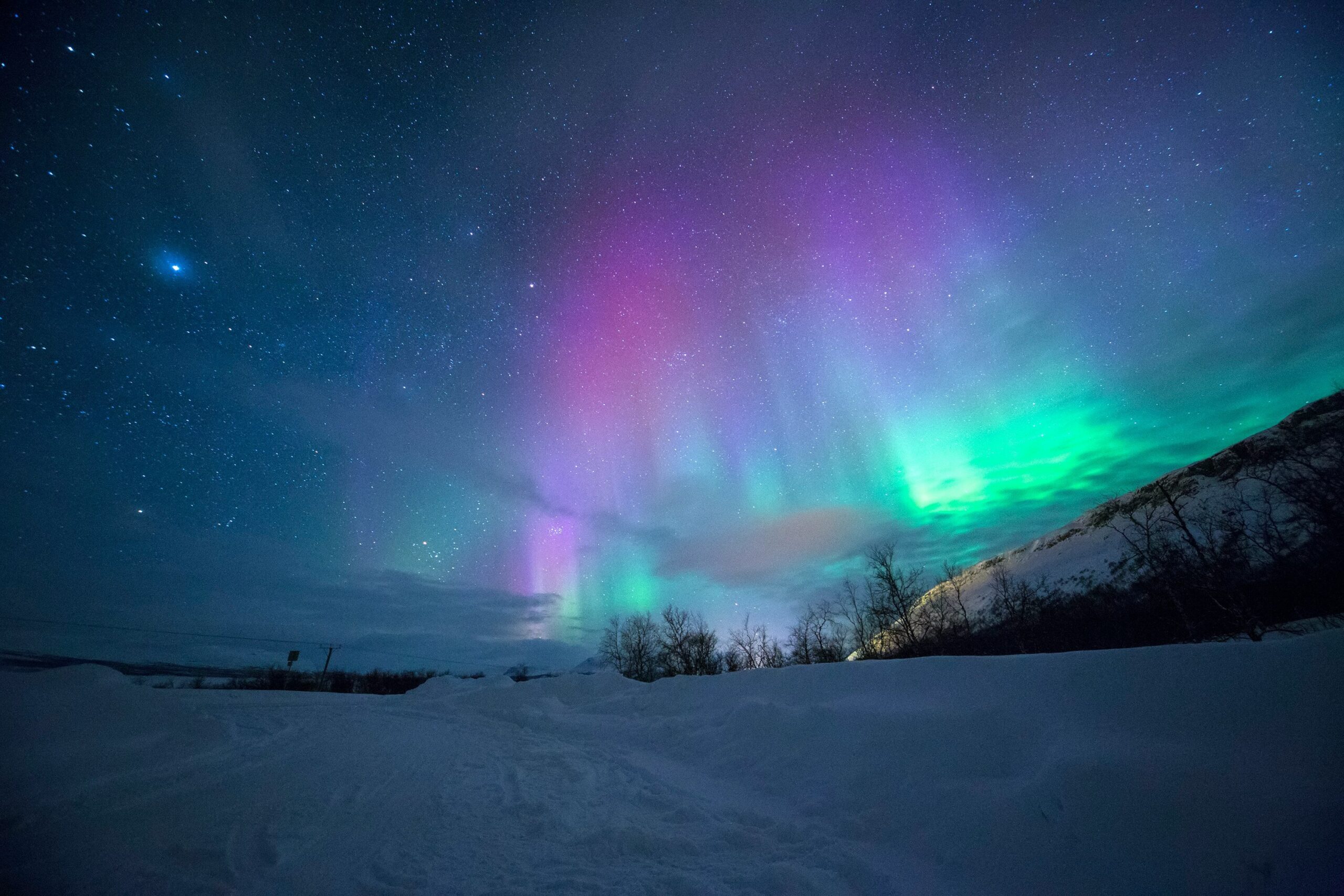 A photo of the northern lights glowing green and pink over a snowy field at night. Credit Lightscape on Unsplash.