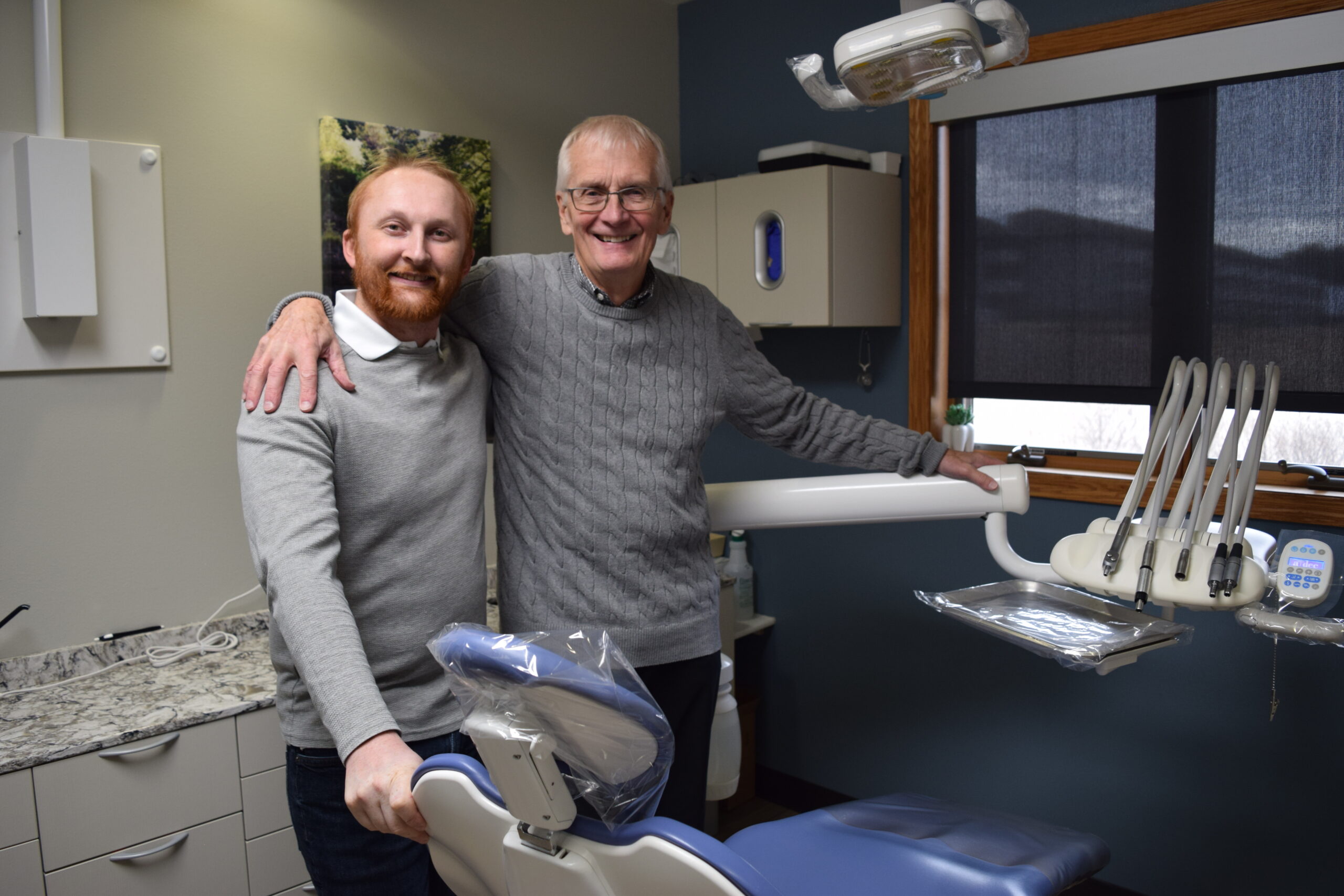 Two men pose for a photo beside an empty dental chair 