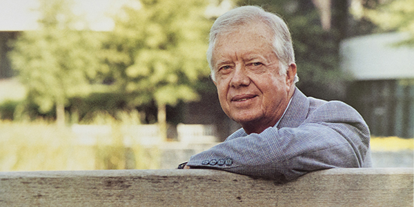 President Jimmy Carter sitting on a bench, smiling and looking at the camera in 1993. He is outside and it is sunny.