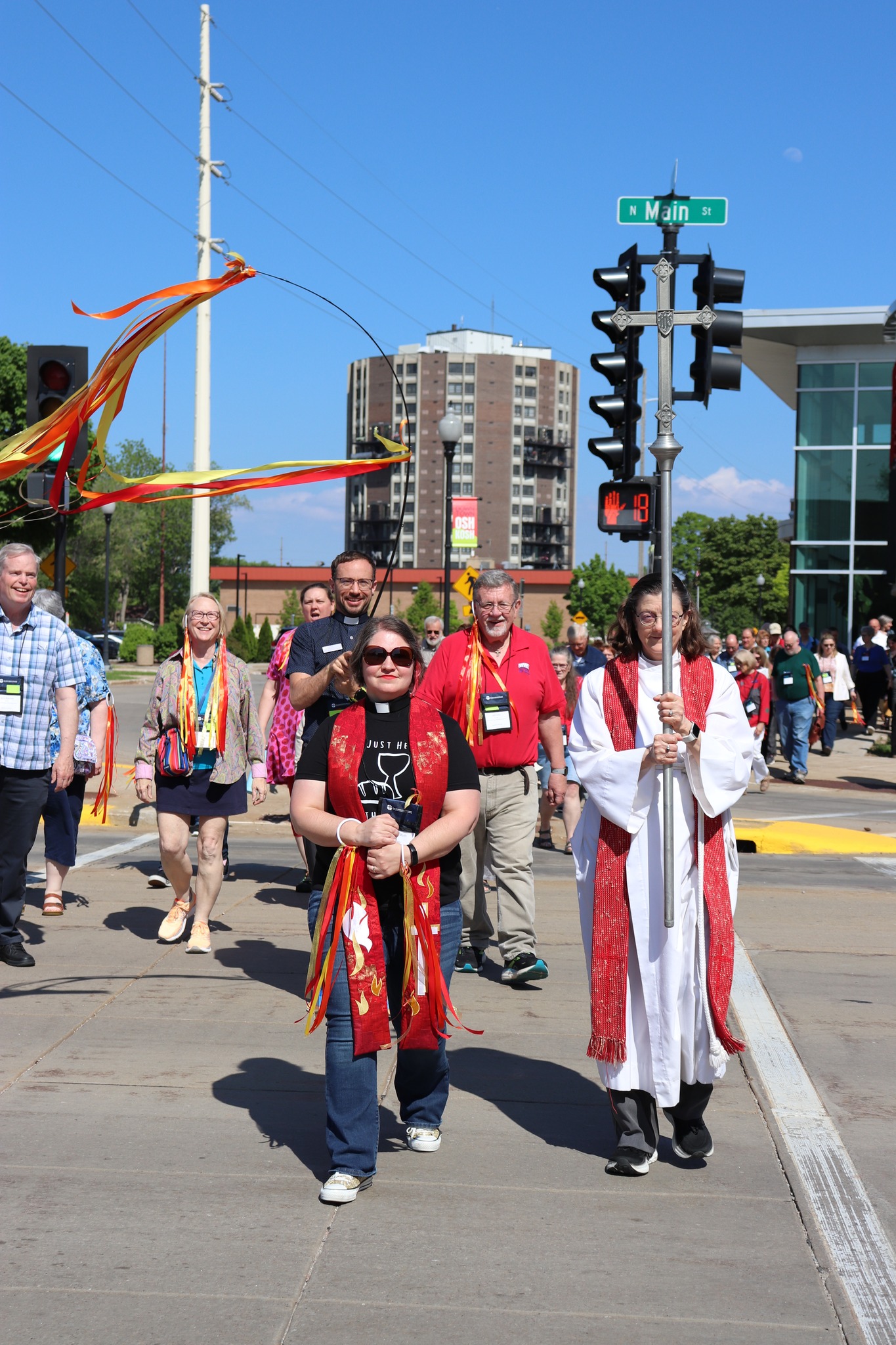 A group of people walking in a procession on a sunny day, led by a man in a white robe holding a tall cross. Colorful ribbons decorate the area.