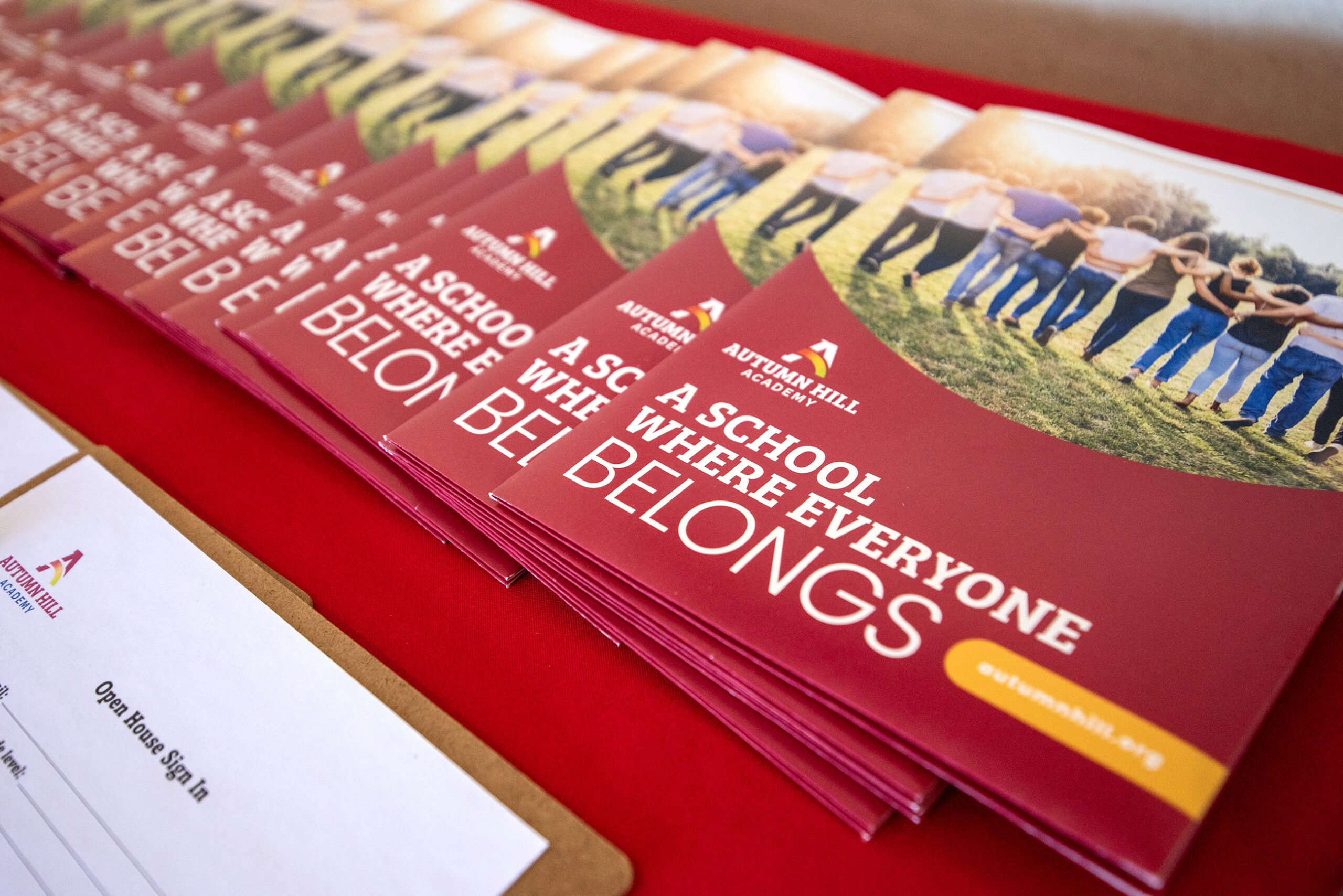 Stack of brochures titled A School Where Everyone Belongs on a red table, next to a clipboard with a sign-in sheet.