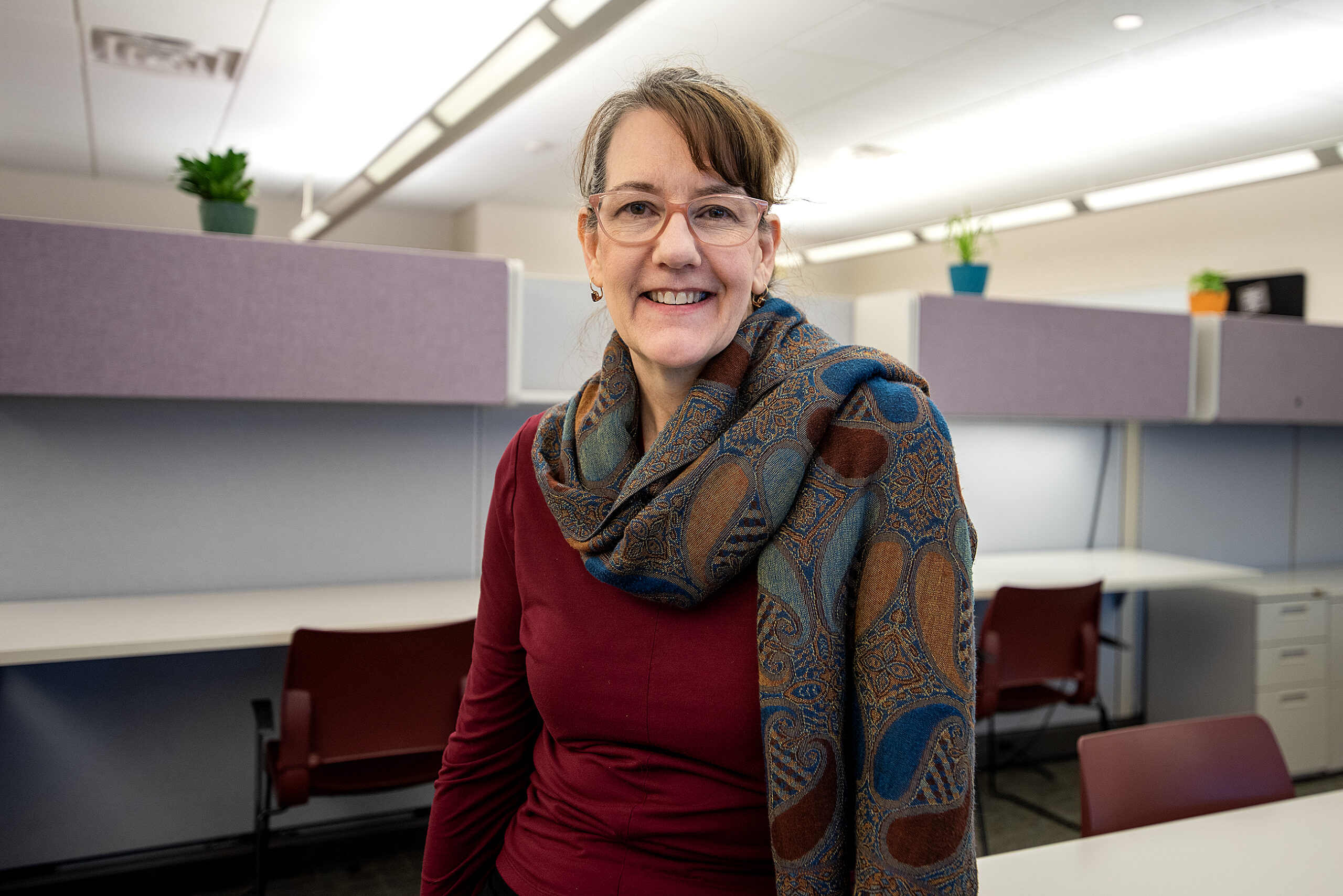 Person with glasses and a patterned scarf smiling in an office setting with desks and chairs.