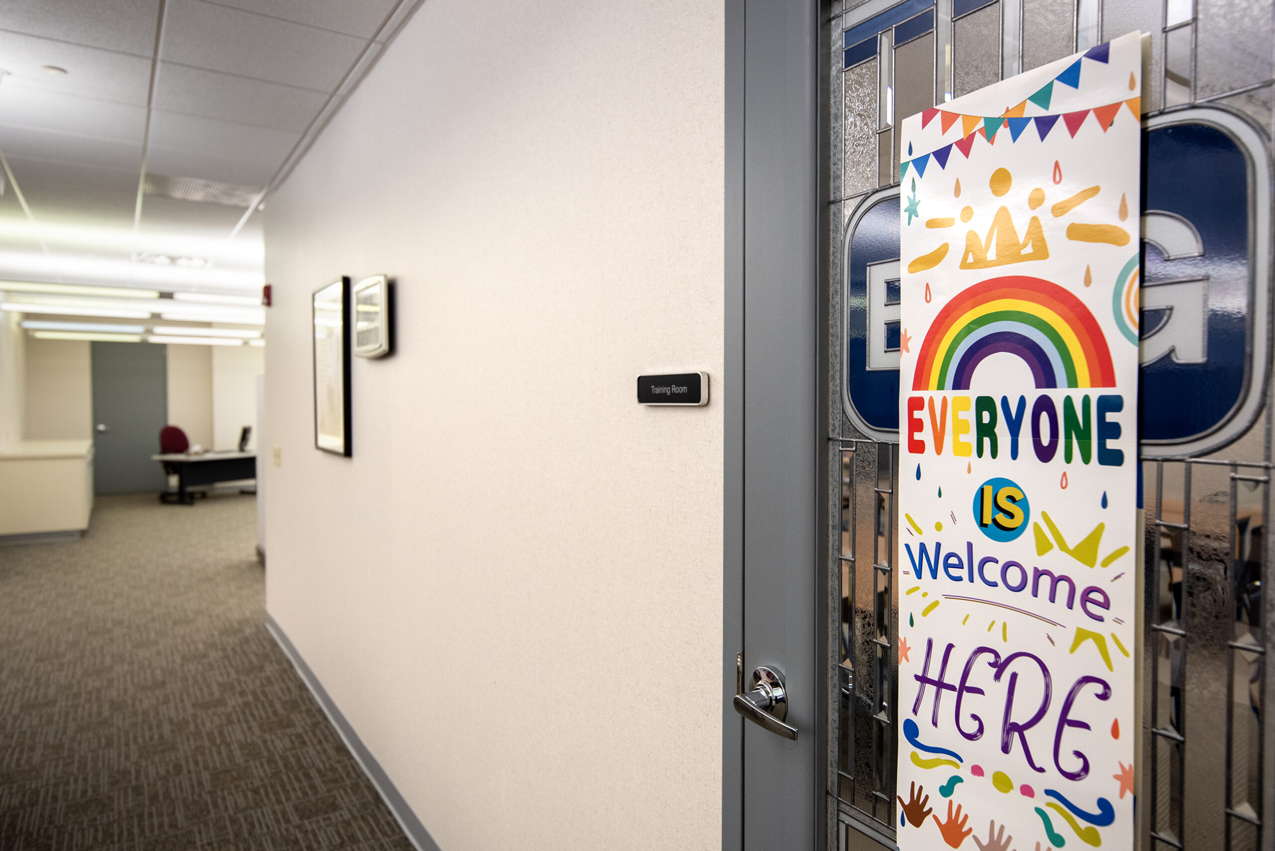 Colorful sign on a door reads Everyone is welcome here with rainbow and star designs. Hallway in the background.