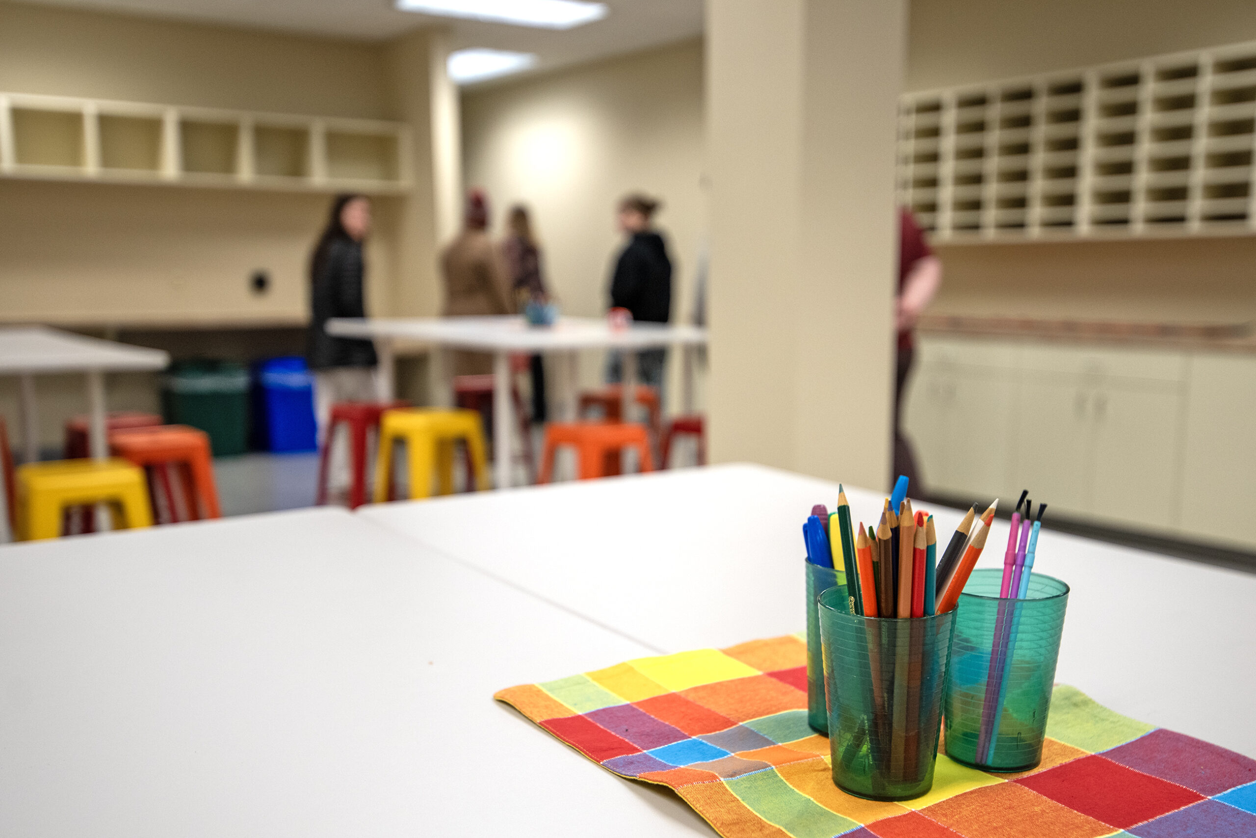 Pencils and a colorful cloth on a table in the foreground, with people conversing near bar stools and tables in a room with beige walls in the background.