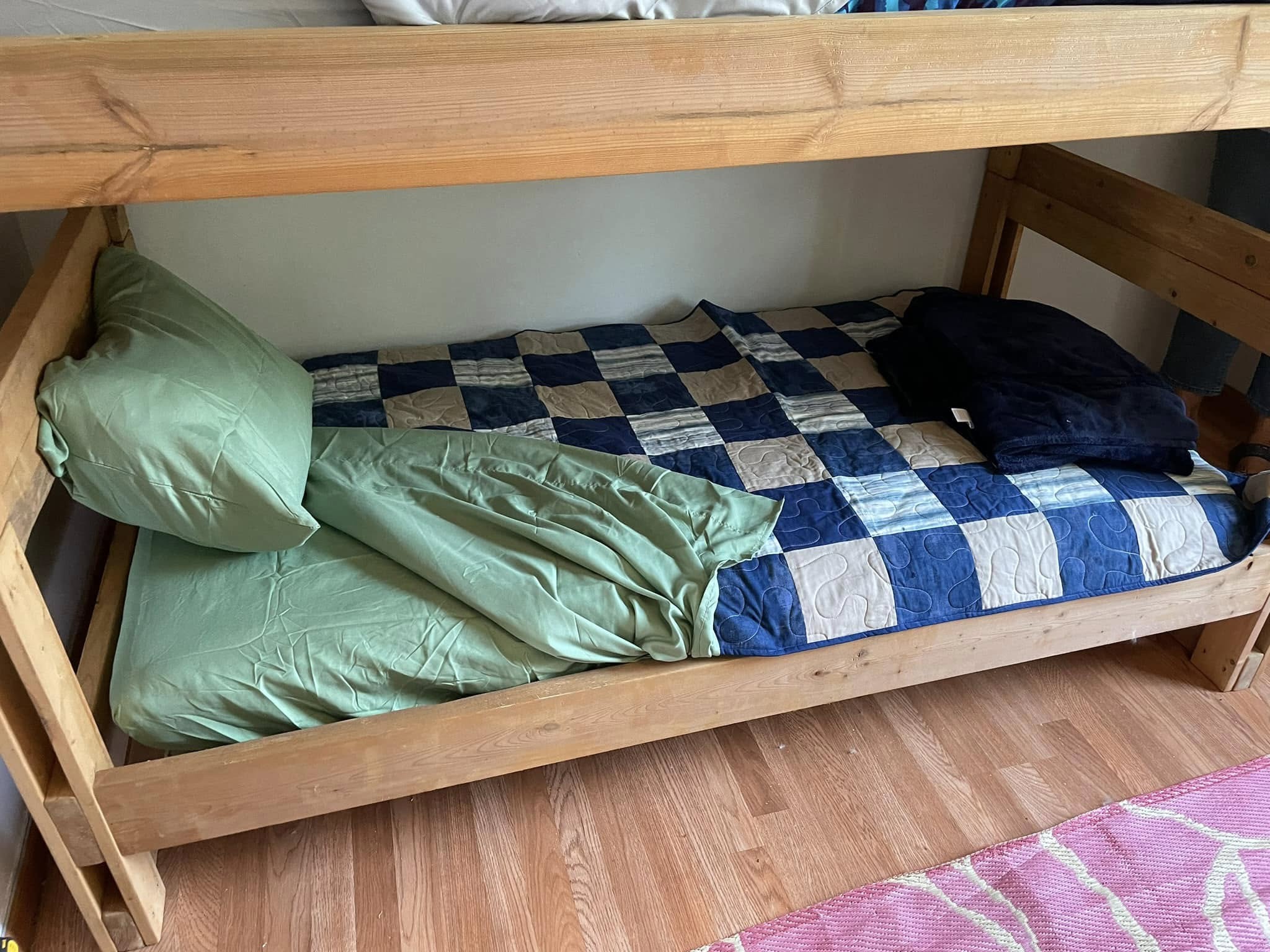 Lower bunk bed with a blue and green checked blanket, green pillow, and crumpled green sheet. A folded dark blue blanket is at the foot of the bed. Wooden frame on wooden floor.