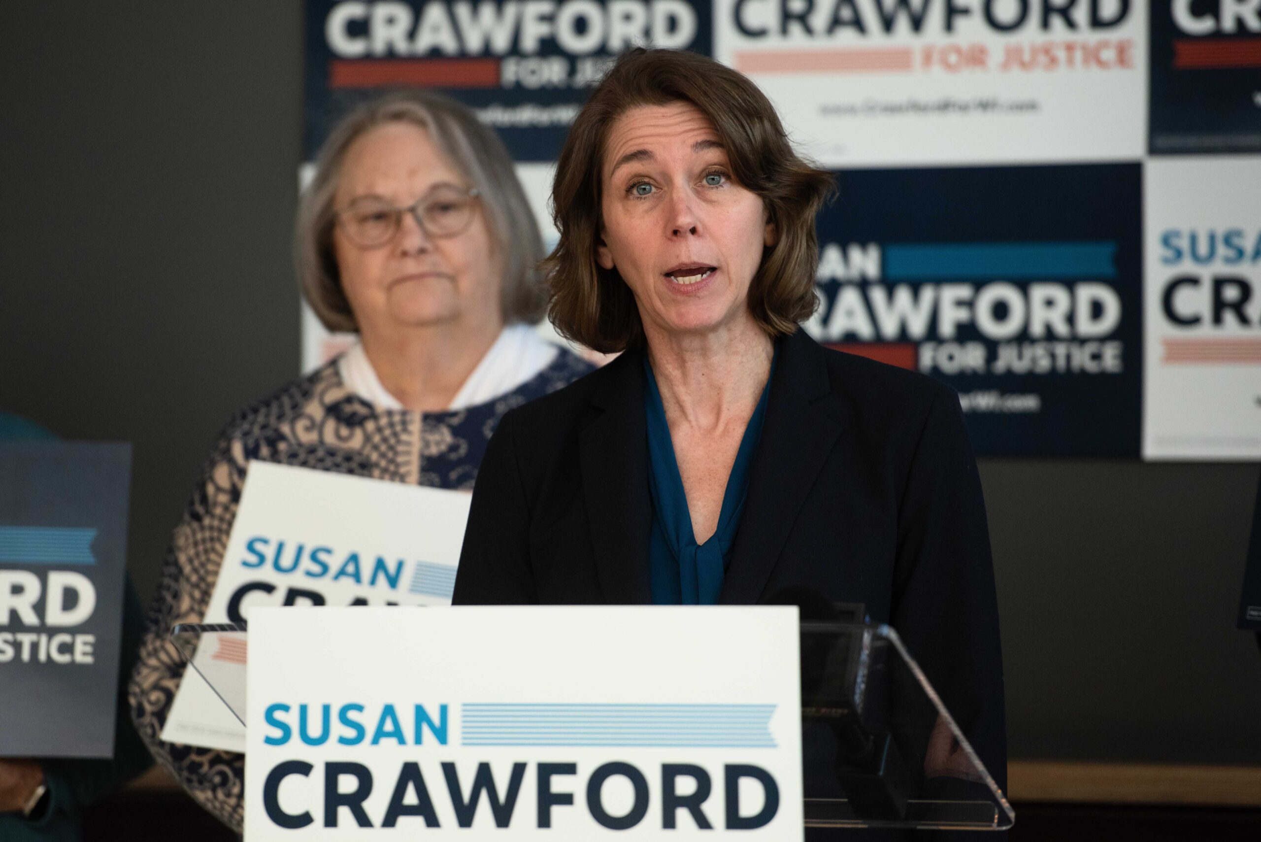 Two women at a podium, one speaking, in front of a backdrop featuring Susan Crawford campaign signs.