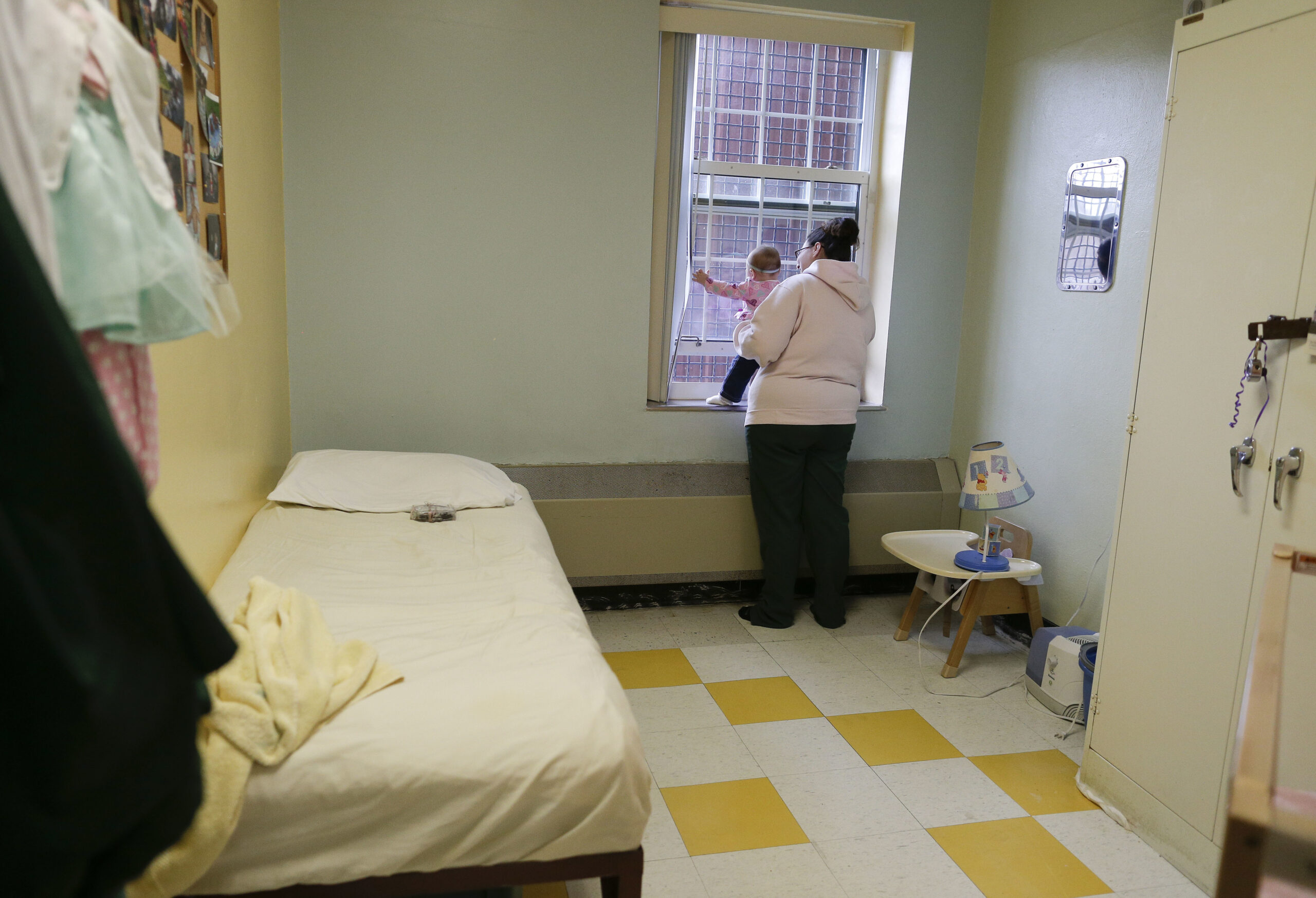 A woman in a white hoodie stands at a barred window holding a child. The room has a bed, small table, lamp, and wardrobe.