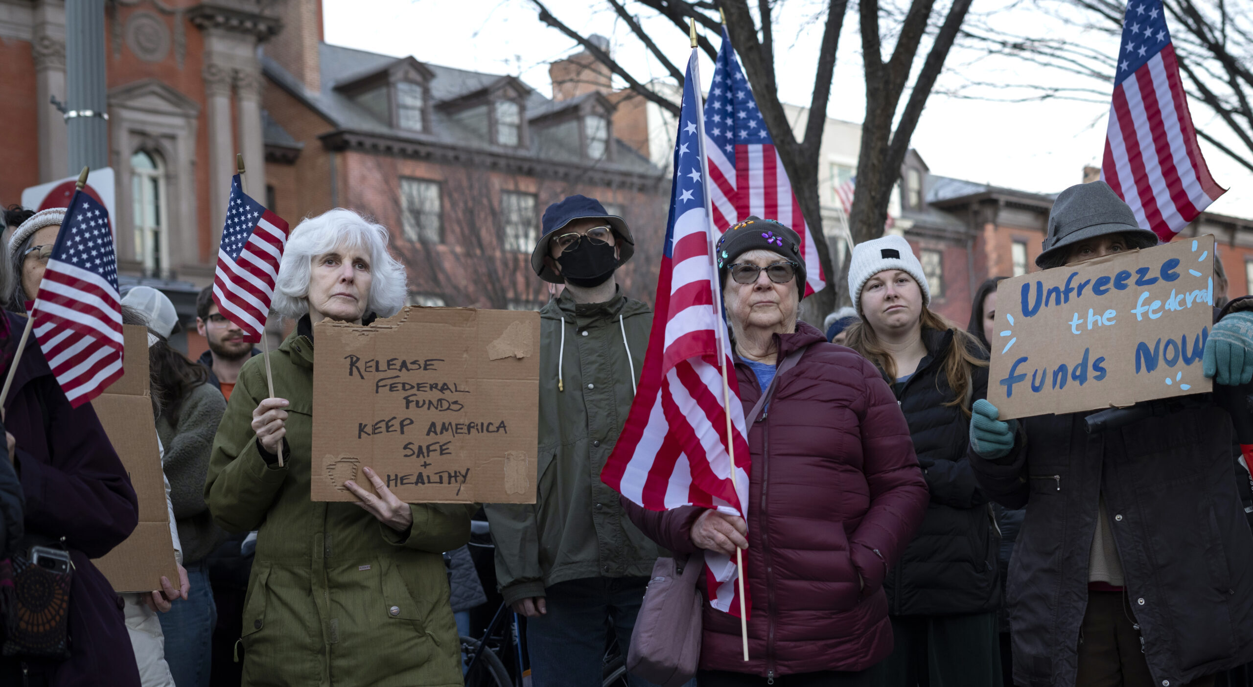 A group of people holding signs and American flags at an outdoor gathering. Some signs read Release Federal Funds and Unfreeze the Funds Now.