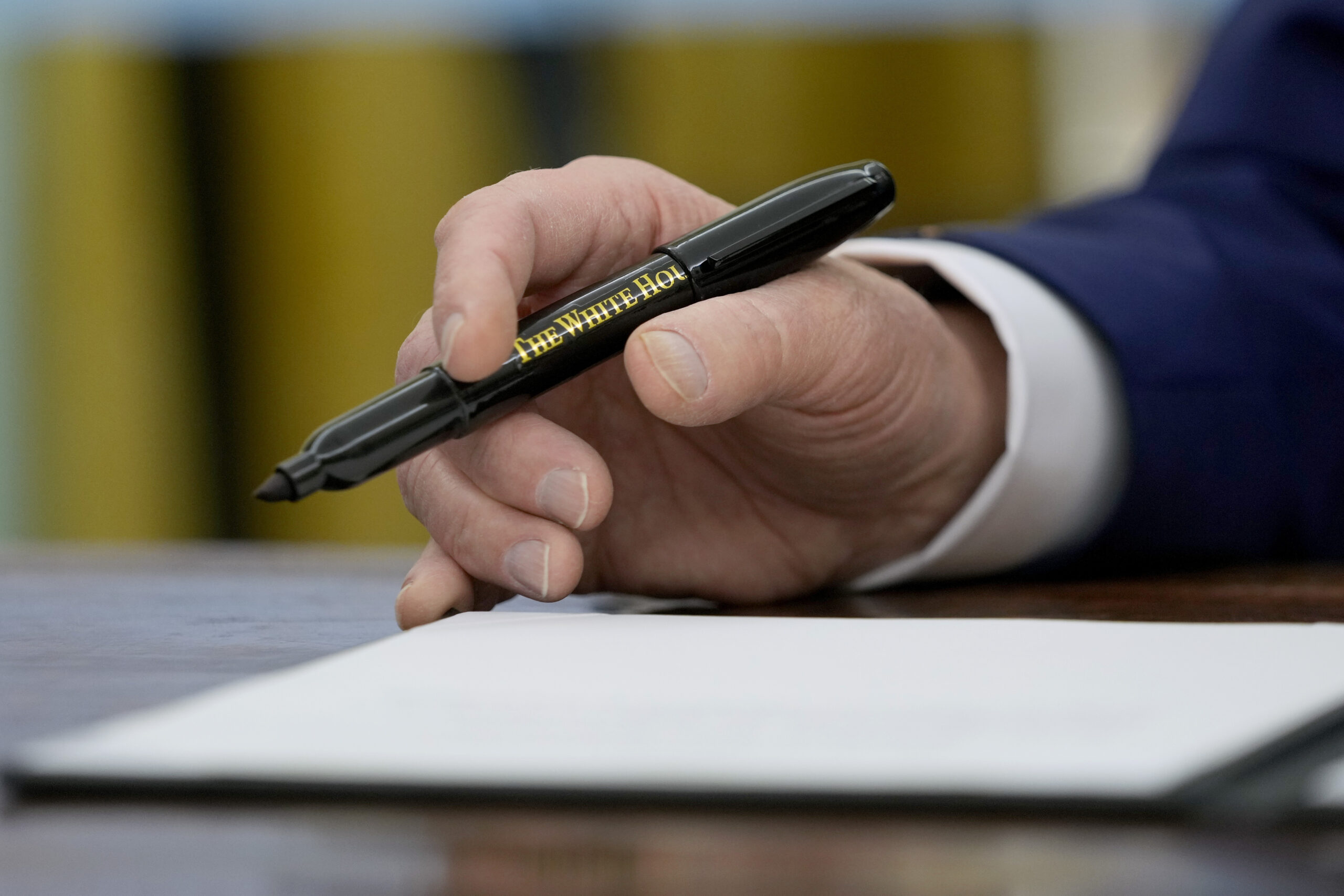 A hand holding a pen labeled The White House poised above a sheet of paper on a table.