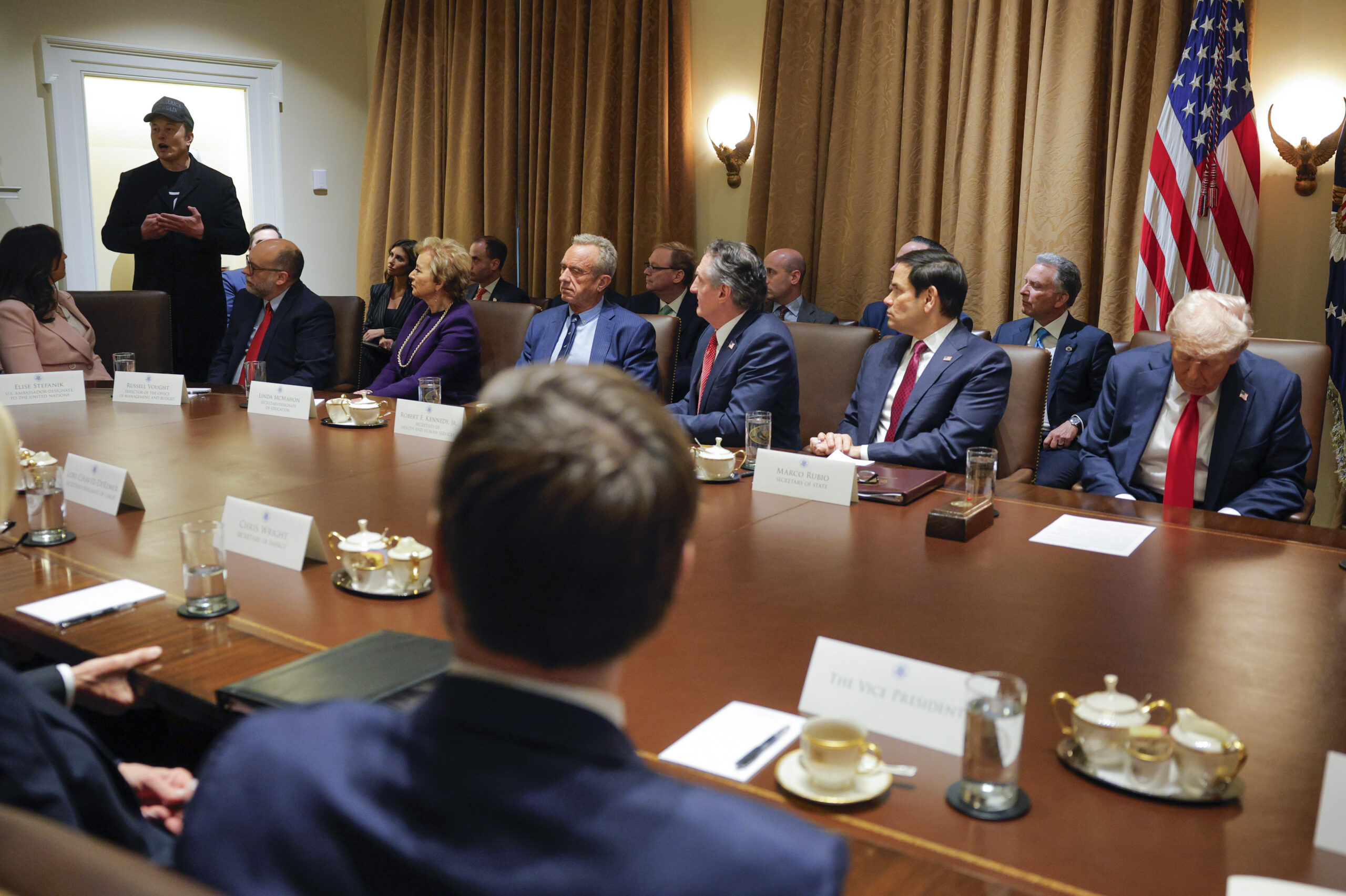 A group of people in formal attire sit around a long conference table during a meeting, with nameplates and microphones on the table. An American flag is visible in the background.