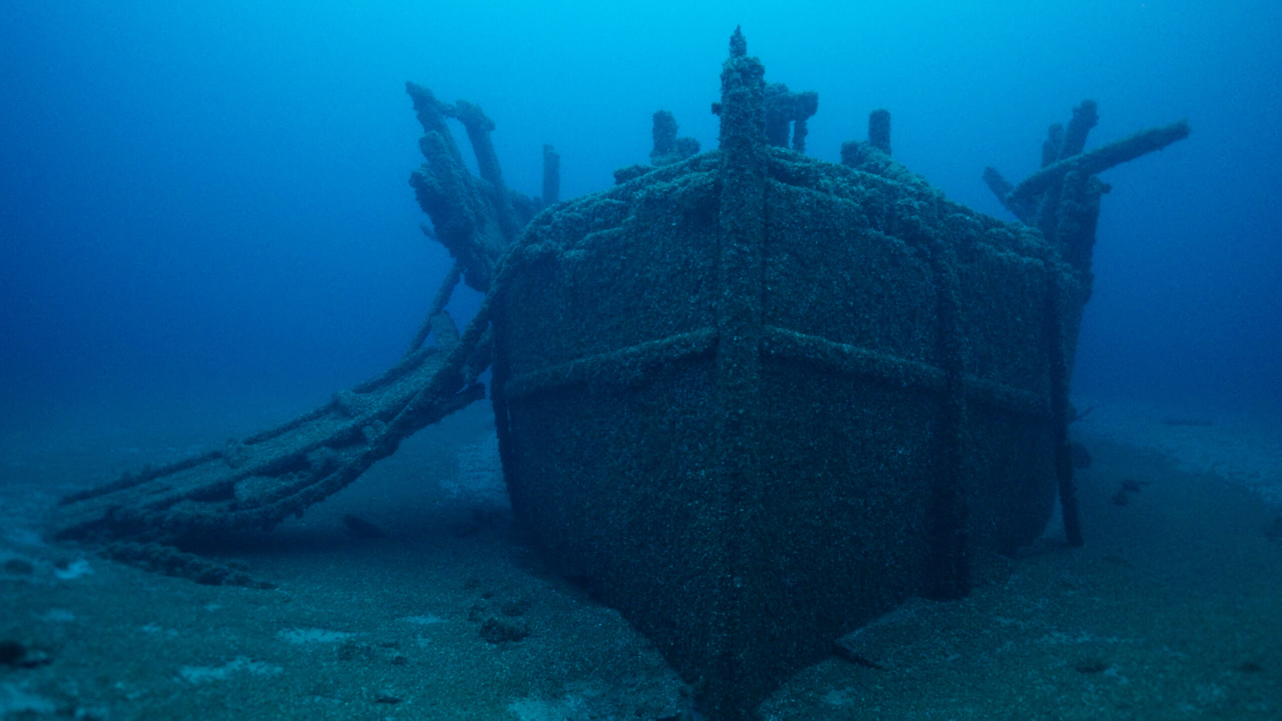 Sunken shipwreck underwater, covered in algae and marine growth, resting on the seafloor, with the bow and part of the wooden structure visible.