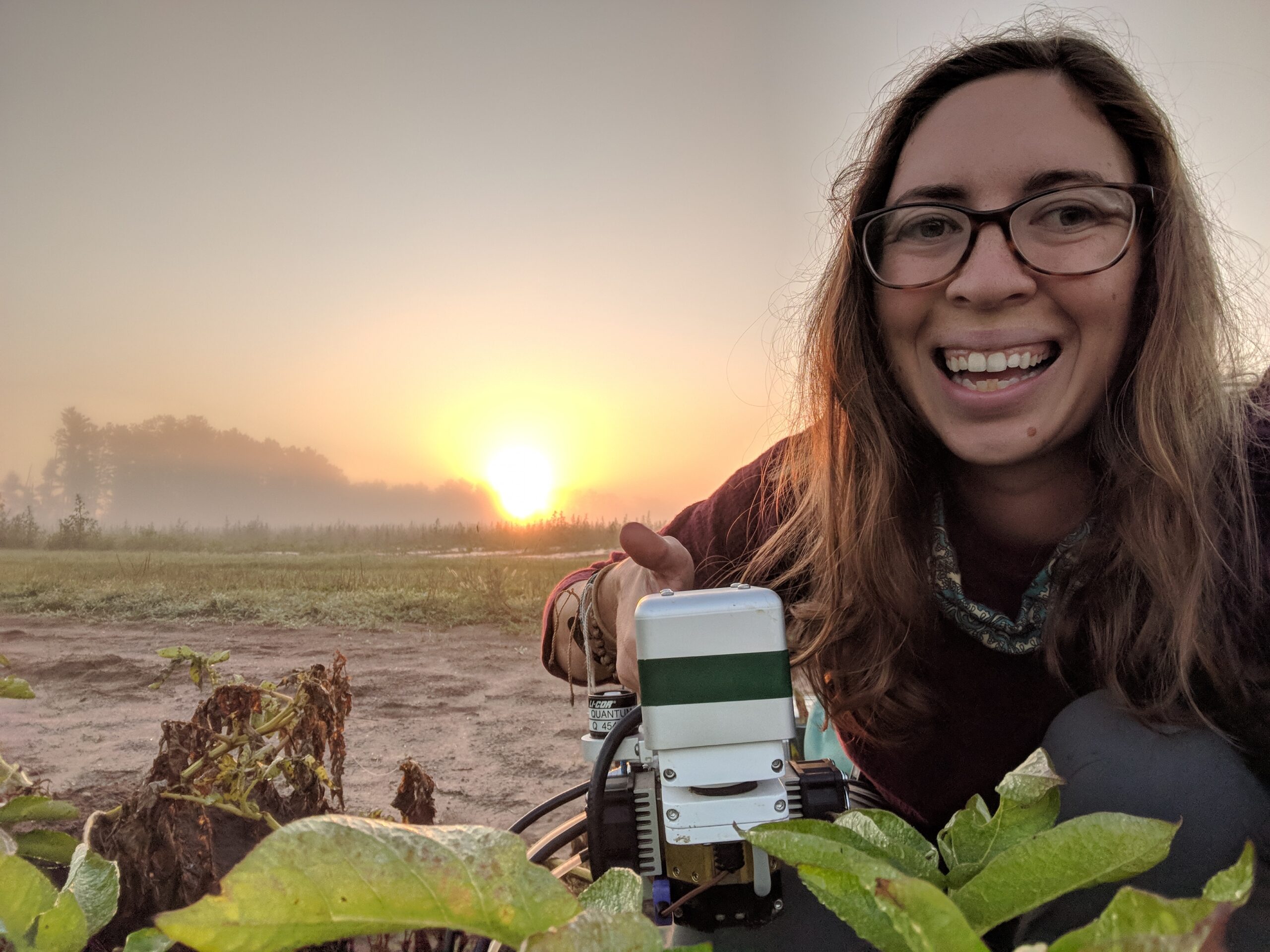 A woman with glasses smiles while using a scientific instrument in a field at sunrise. She is surrounded by plants and the sun glows in the misty background.