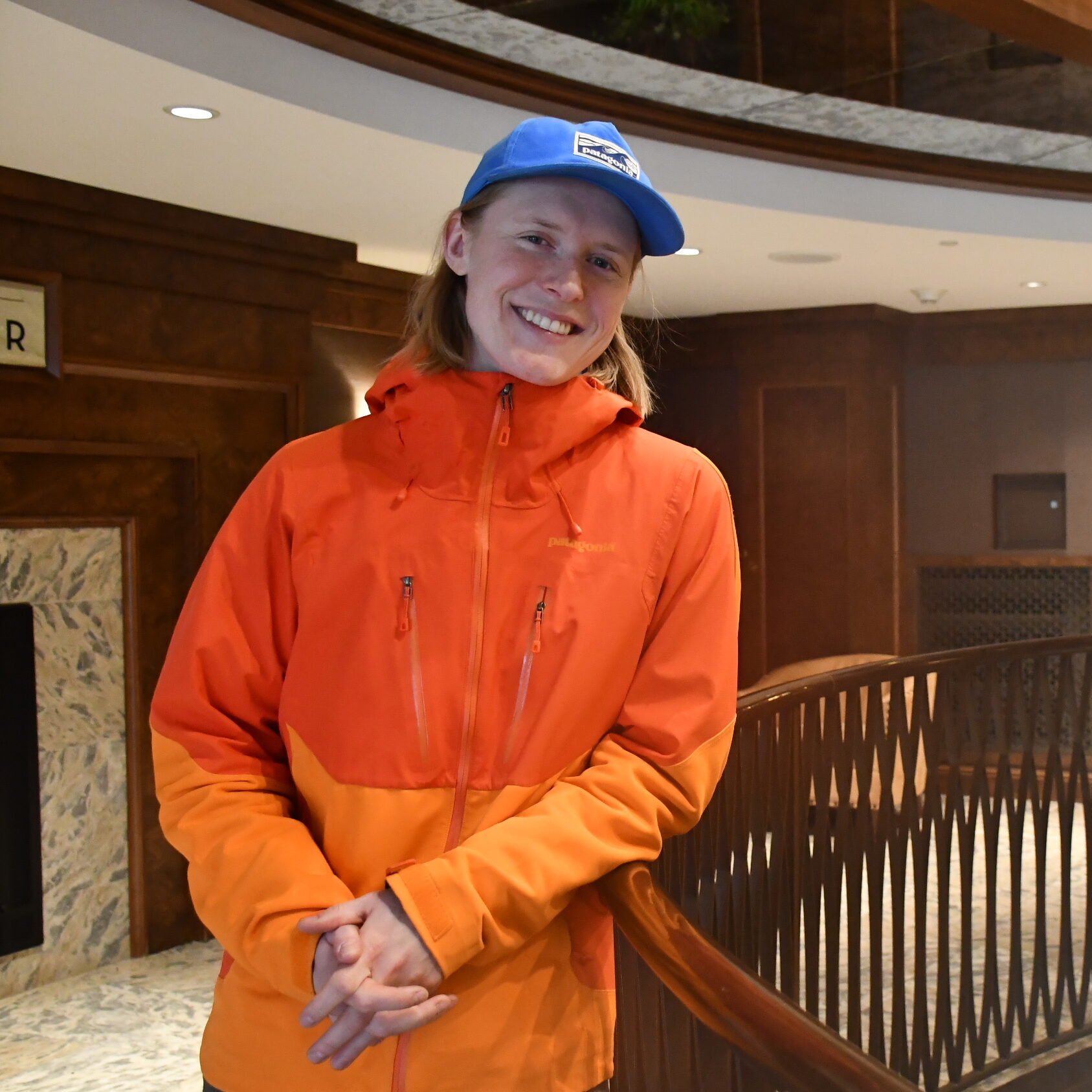 Person in an orange jacket and blue cap stands in a hotel lobby near a railing, looking at the camera.