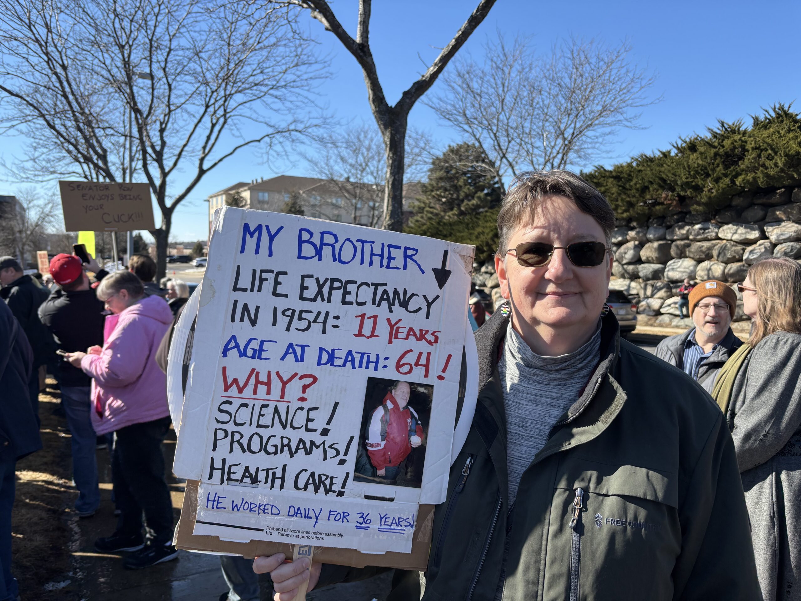 A person holds a sign about their brothers life expectancy at a protest. People are gathered in the background.