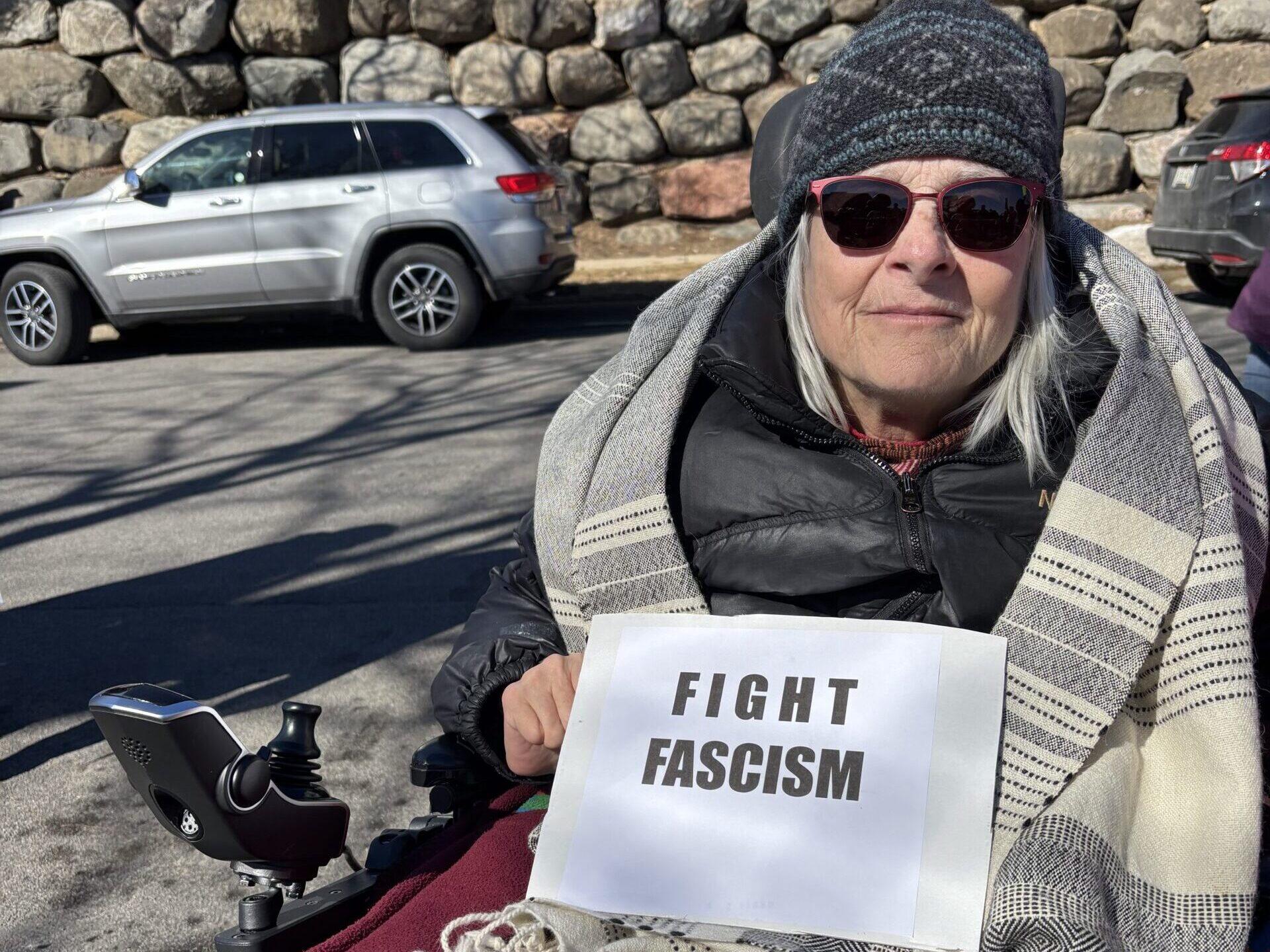 A person in sunglasses and winter attire sits in a wheelchair, holding a sign that reads FIGHT FASCISM on a sunny day near parked cars and a stone wall.