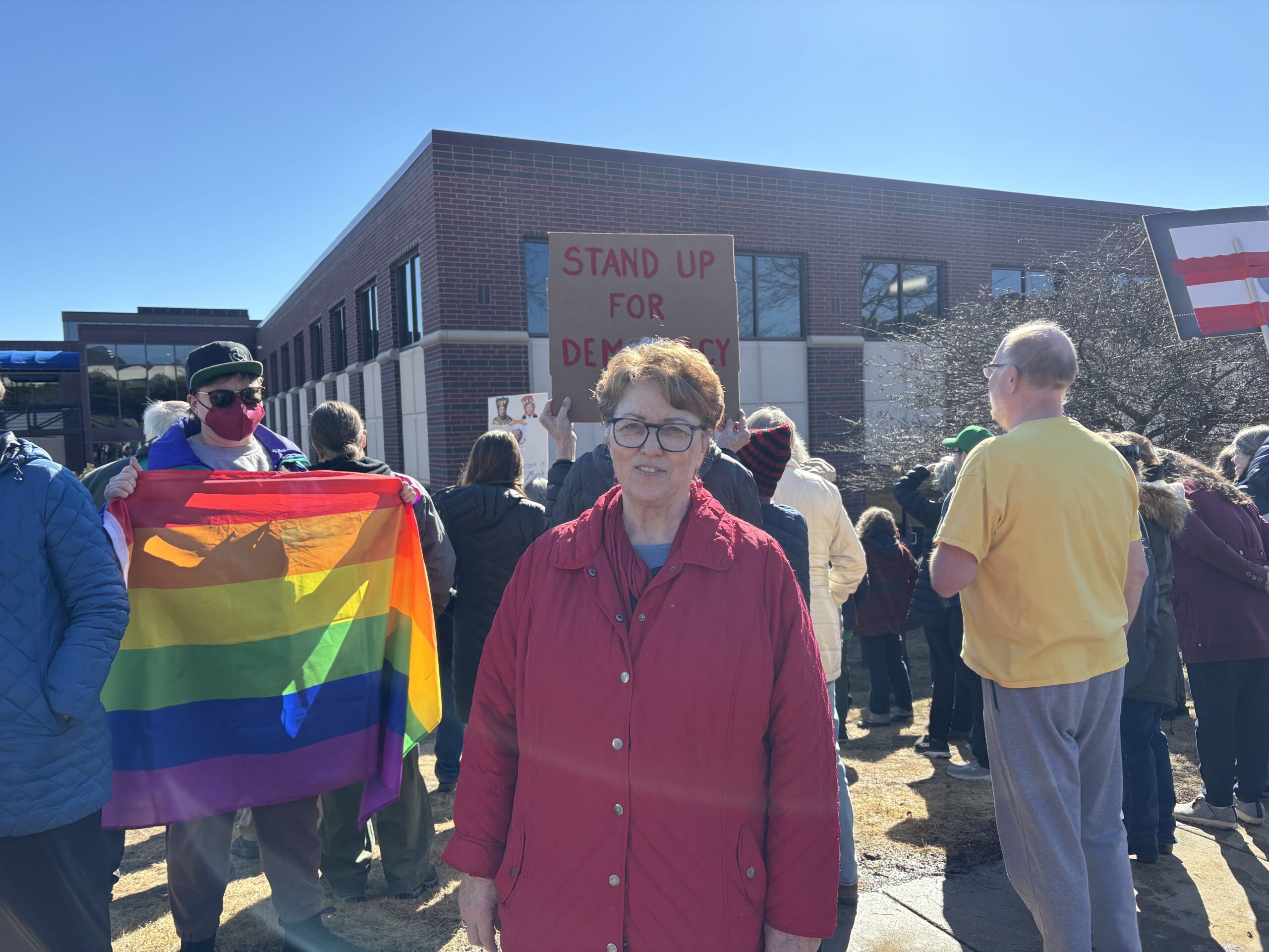 A person in a red jacket stands in front of a crowd holding a Stand Up for Democracy sign. Someone nearby holds a rainbow flag.