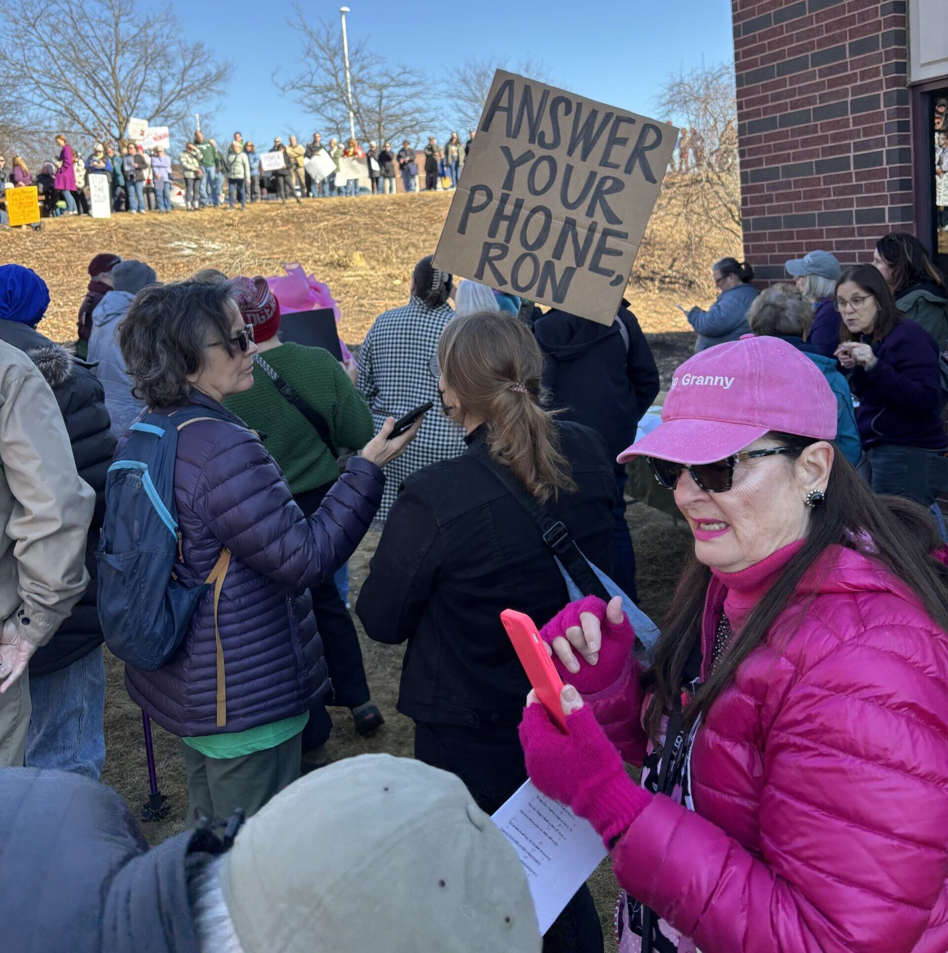 A crowd gathers outdoors holding signs, including one that reads ANSWER YOUR PHONE RON. One person in a pink hat uses a smartphone.