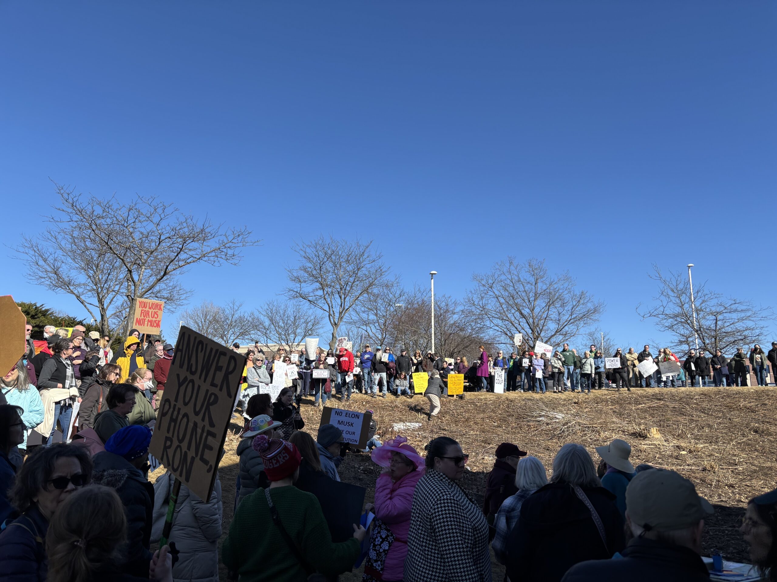 A large crowd stands outdoors holding various protest signs on a sunny day, with leafless trees and a clear blue sky in the background.