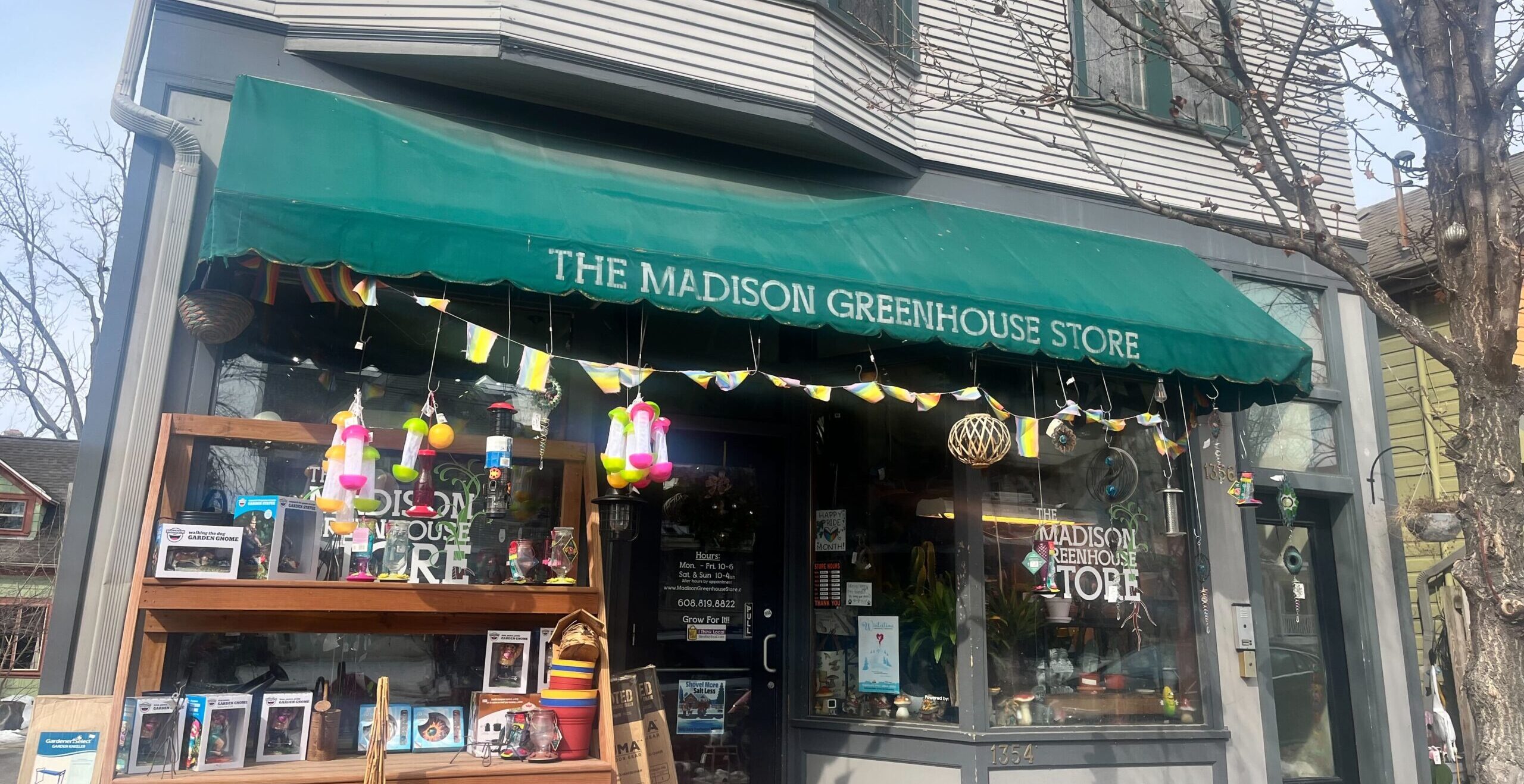 A storefront with a green awning reading The Madison Greenhouse Store. Decorative items hang outside, and various products are displayed in the windows and on shelves.