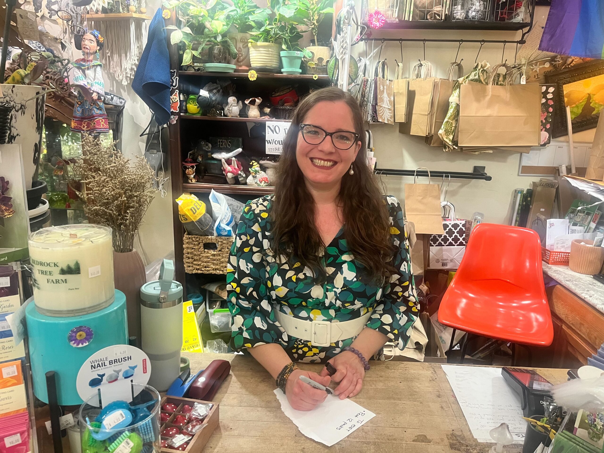 A woman with long brown hair and glasses stands behind a cluttered shop counter, holding a small piece of paper and smiling. Various items and paper bags are displayed around her.