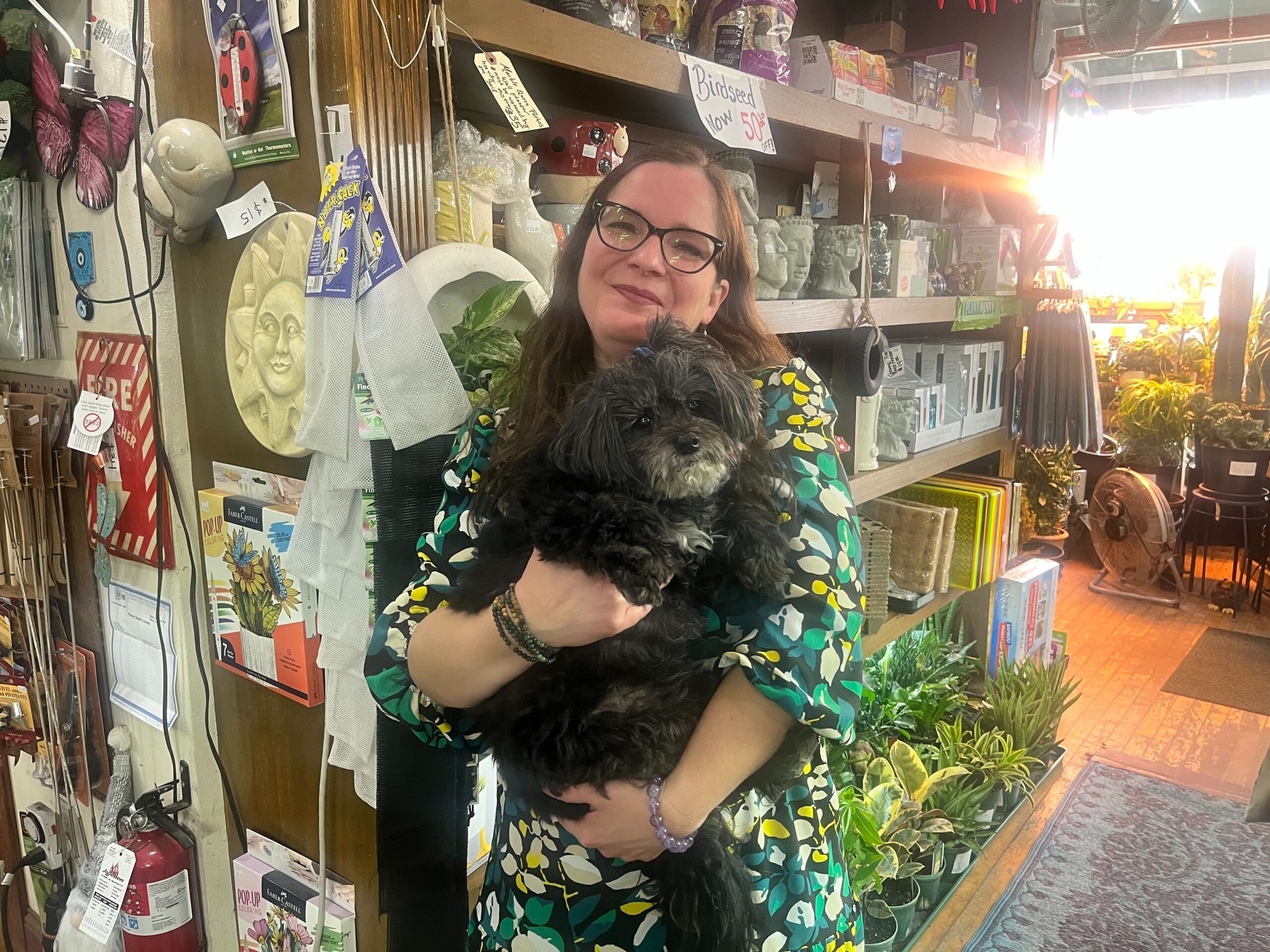 A woman in a floral dress holds a small black dog inside a store filled with various decorative items and plants.