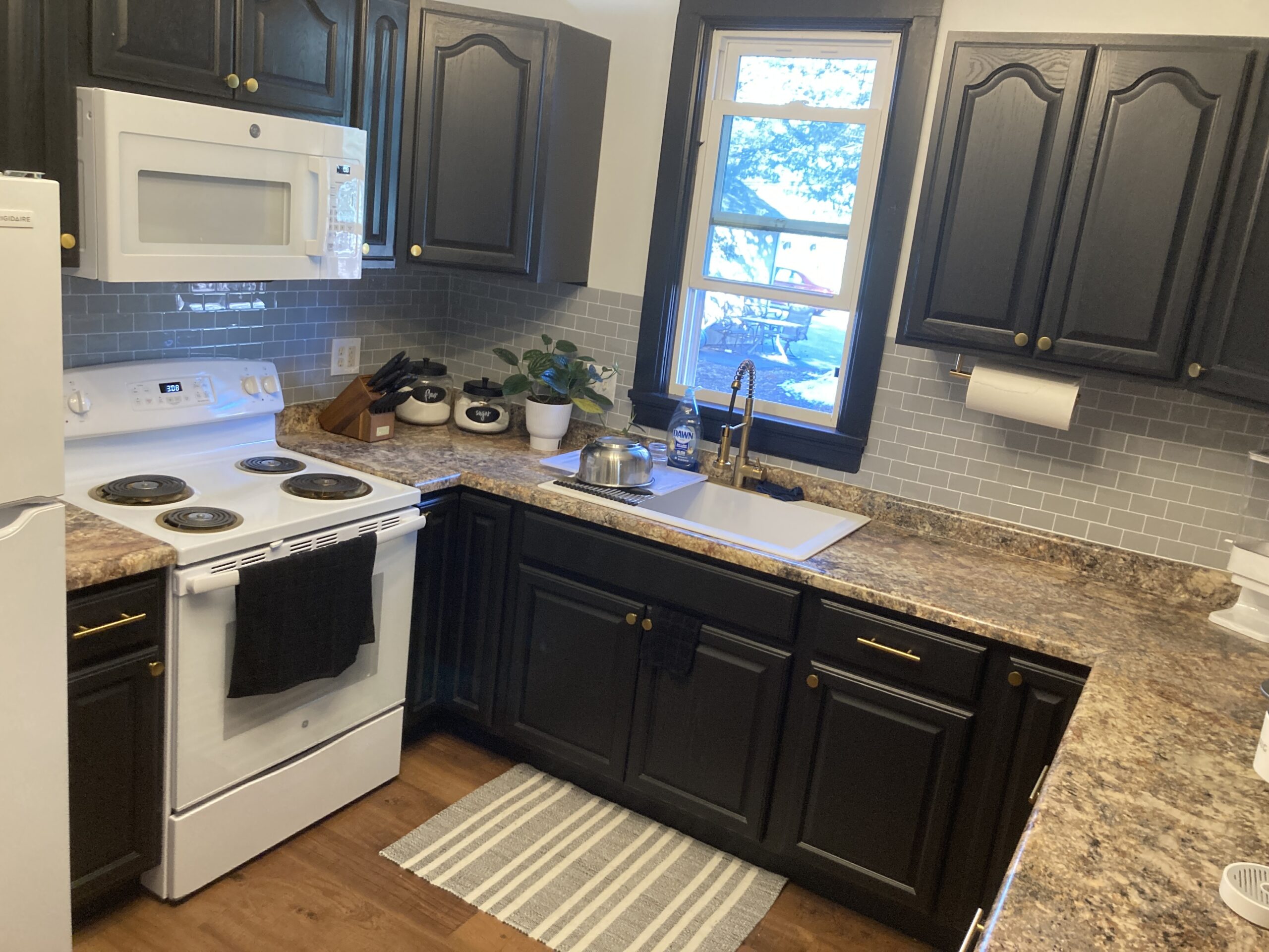 A kitchen with dark cabinets, a white stove and microwave, a brown countertop, and a sink under a window. A small plant is on the counter next to a teapot. A striped rug lies on the floor.