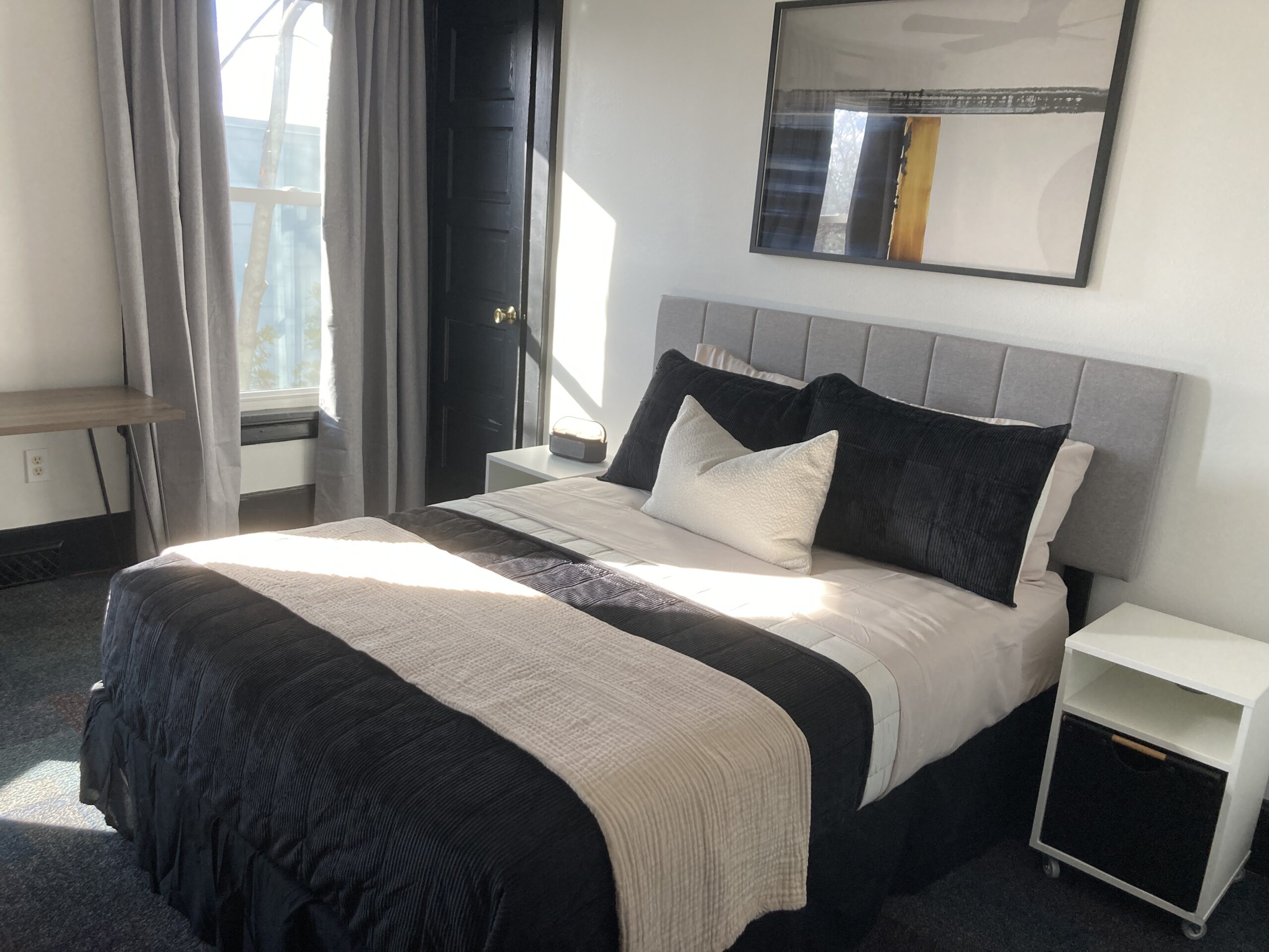 Bedroom with a neatly made bed featuring black and white bedding, two nightstands, a desk, a window with curtains, and a framed mirror on the wall.