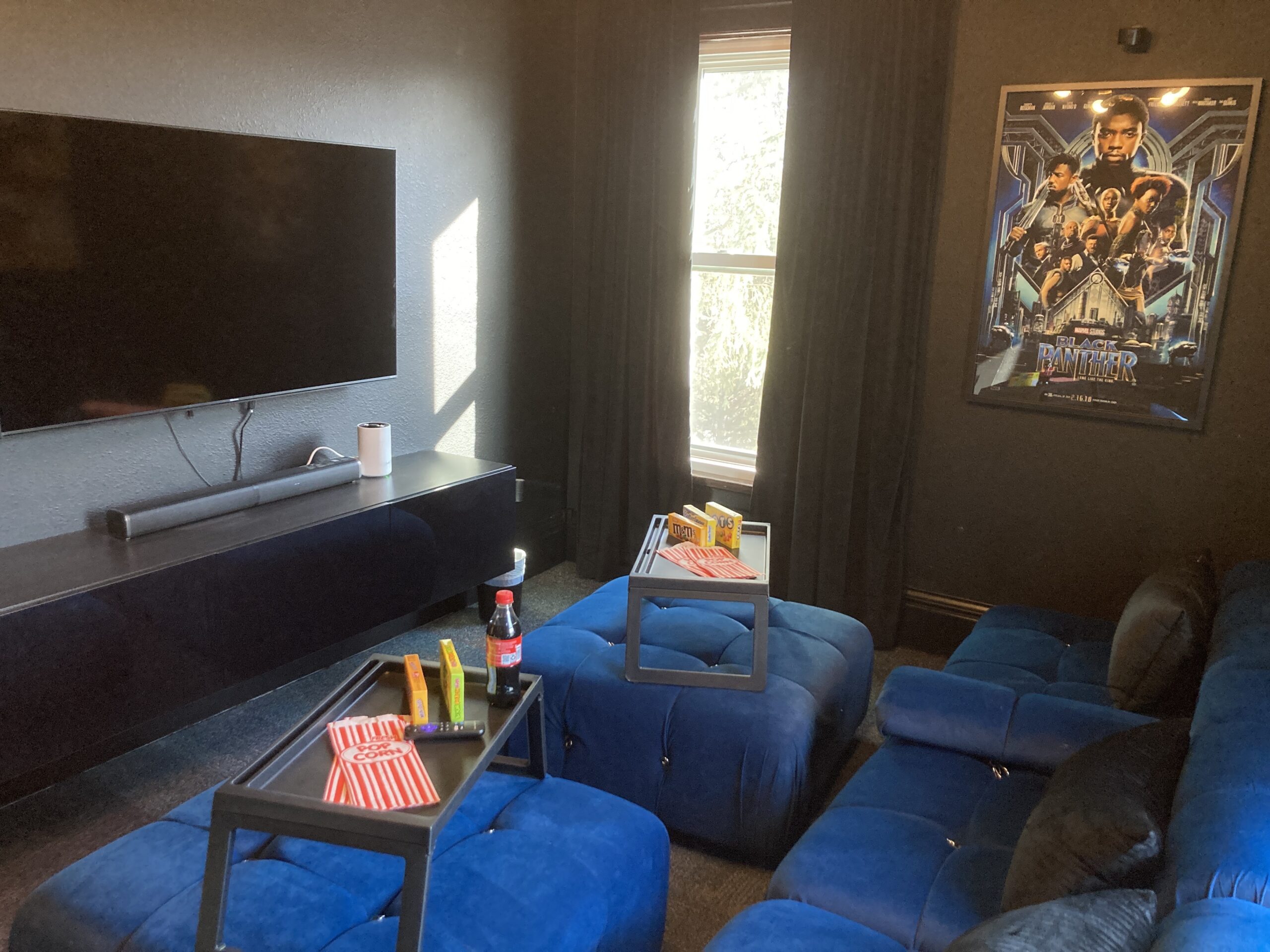 A home theater room with a large TV, blue sectional seating, a Black Panther poster on the wall, and snack trays holding drinks and popcorn.