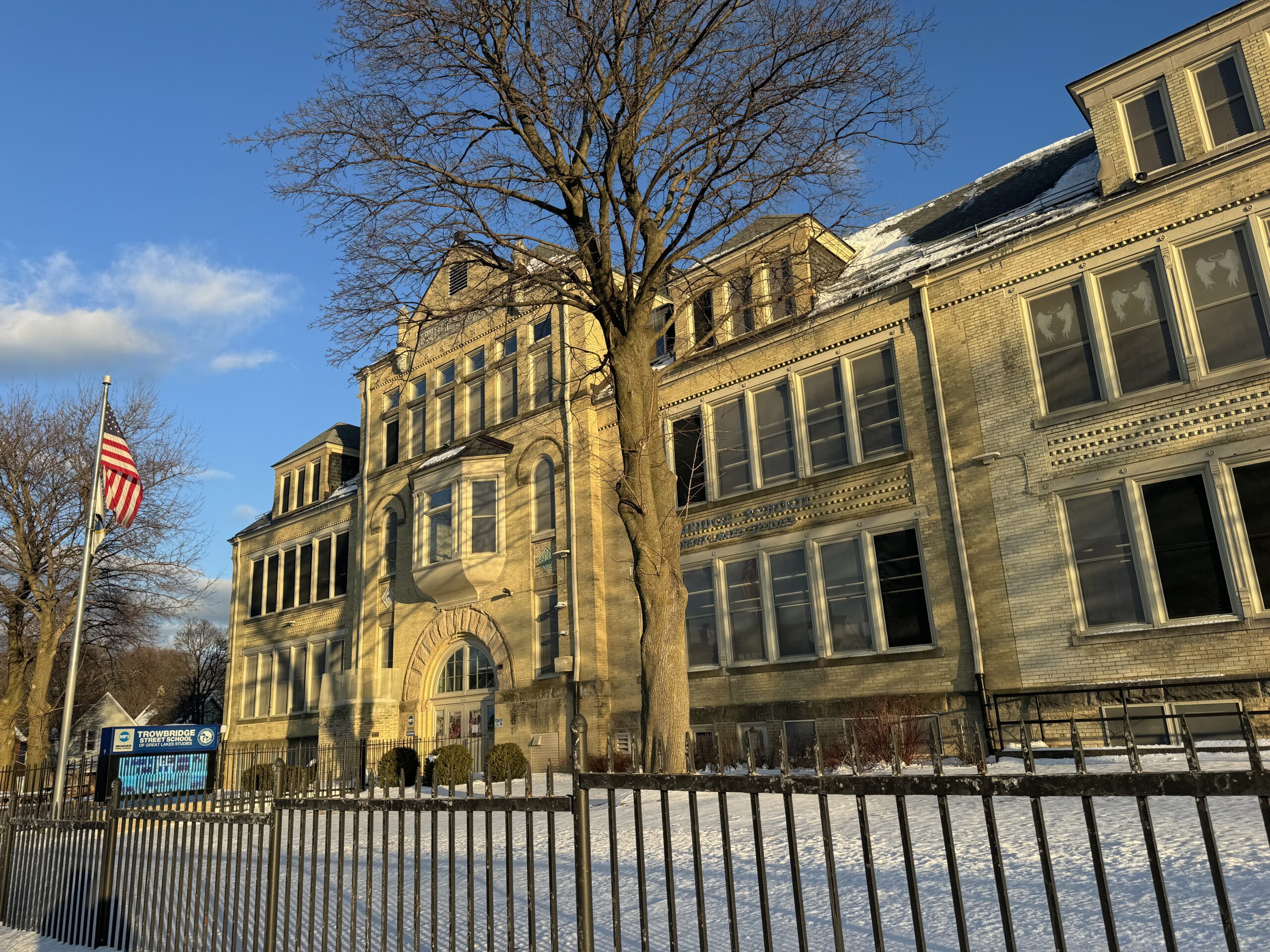 A tall tree and American flag in front of a large, historic brick building, partially covered with snow, under a clear blue sky.