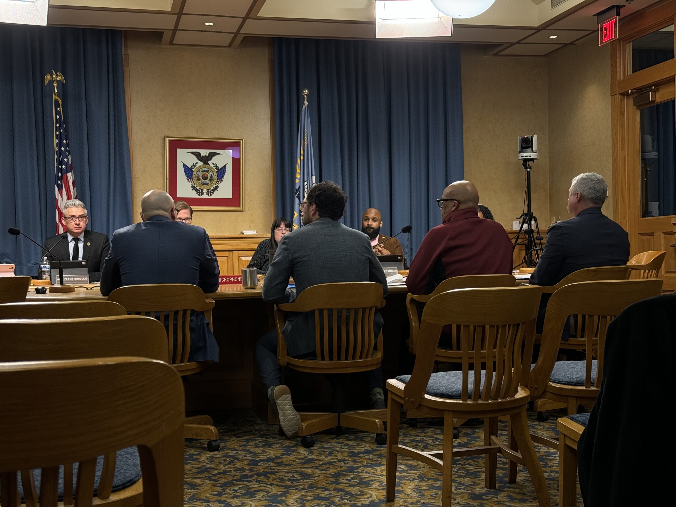 People sitting around a conference table in a meeting room, with flags and a framed emblem on the wall in the background.