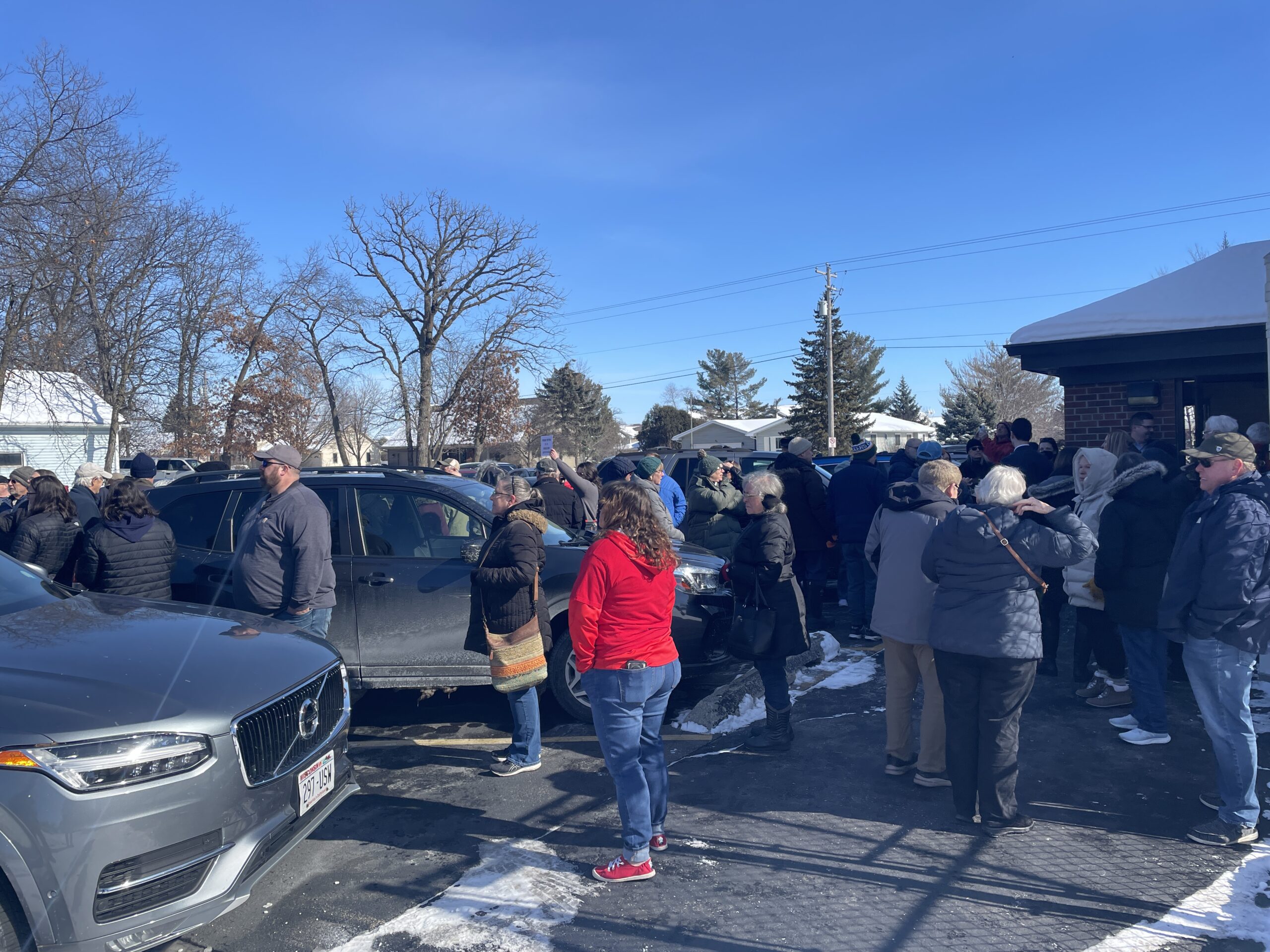 A crowd of people, bundled in winter clothing, stands outside on a snowy sidewalk next to parked cars under a clear blue sky.