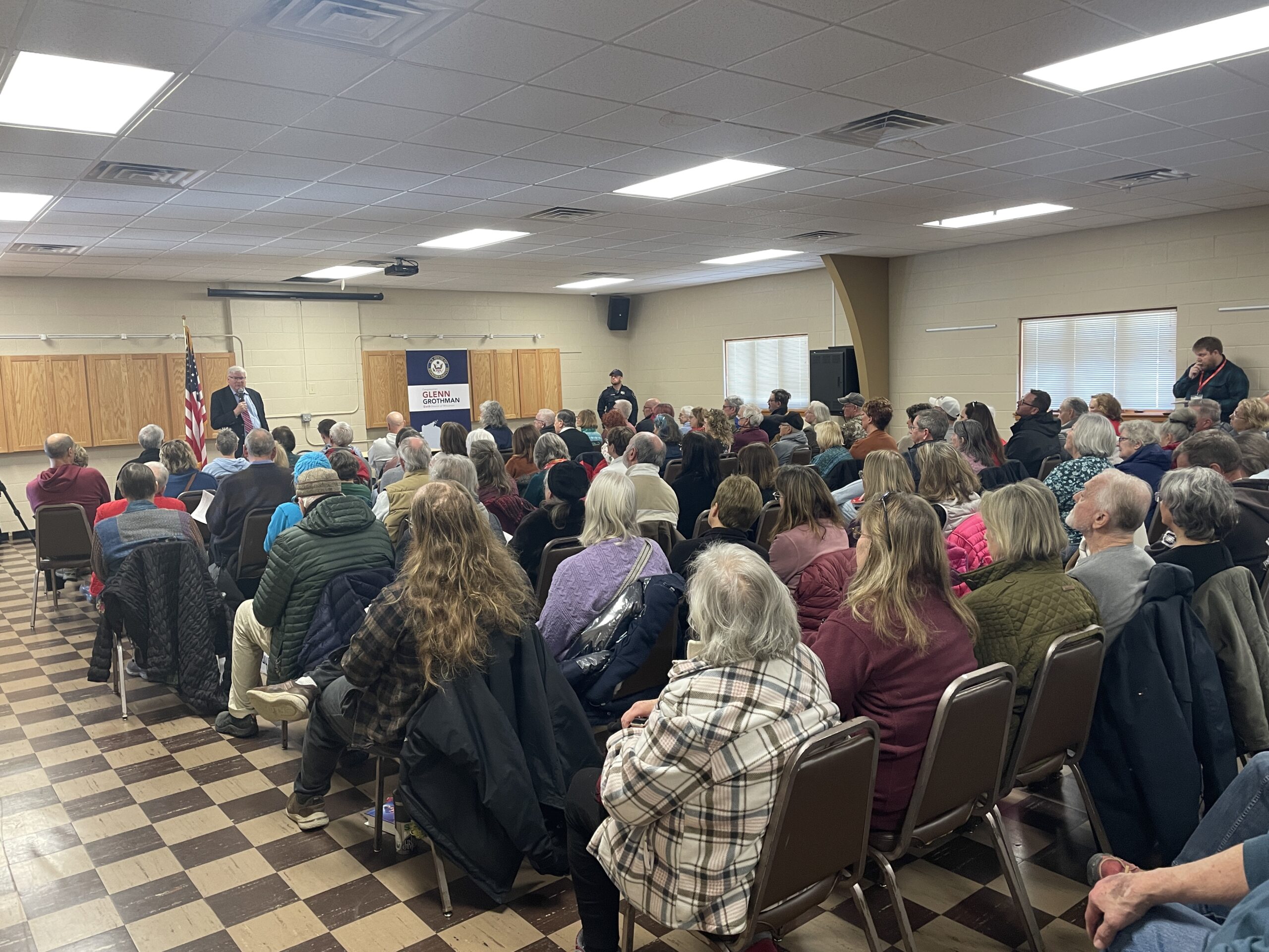 A person speaks at a podium to a seated audience in a community hall. An American flag is displayed nearby.